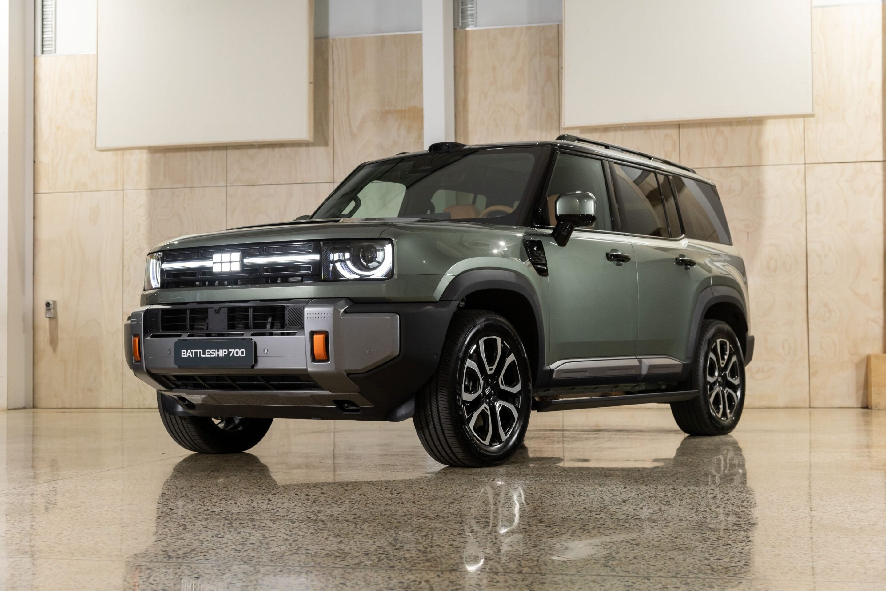 Front three-quarter view of a green SUV in a showroom, displaying a bold grille and round headlights (Battleship 700 on the license plate).