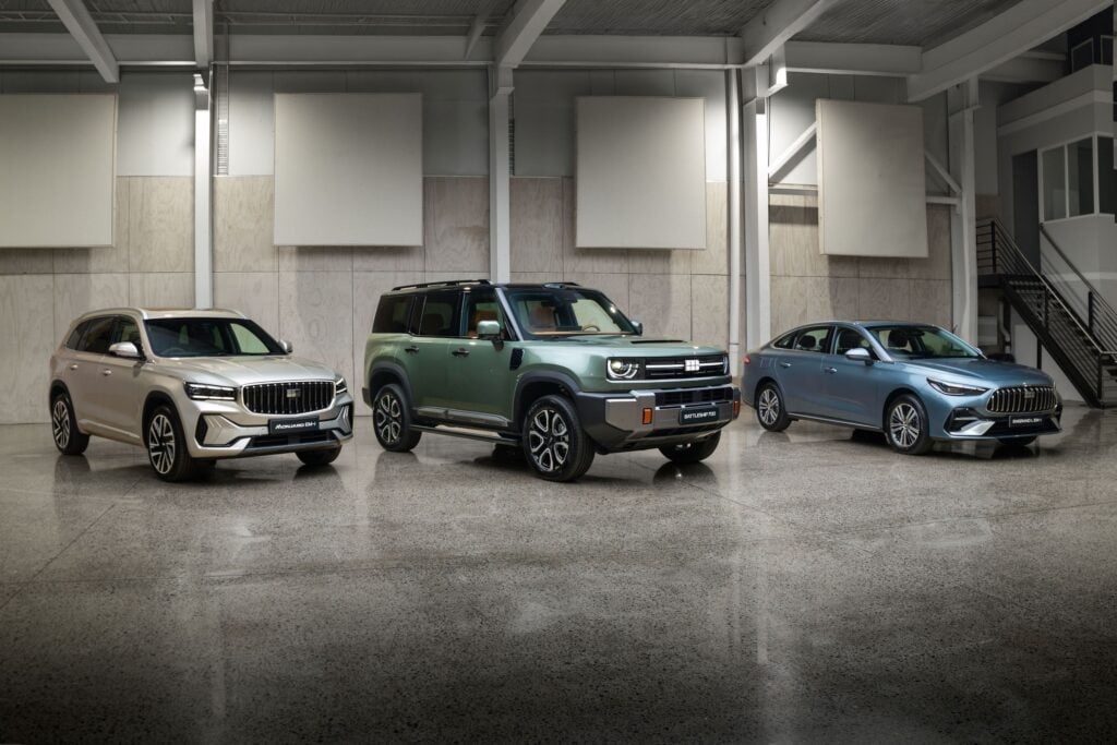 Three new vehicles on display in a showroom: beige SUV on the left, green SUV in the middle, blue sedan on the right, with large panels on the wall behind.