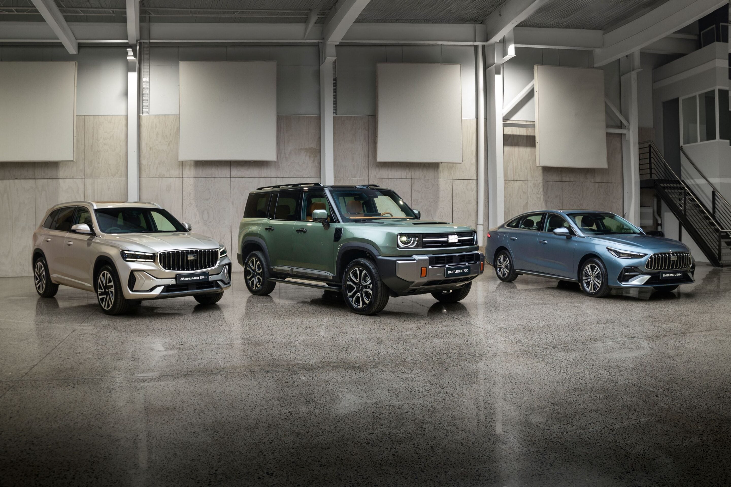 Three new vehicles on display in a showroom: beige SUV on the left, green SUV in the middle, blue sedan on the right, with large panels on the wall behind.