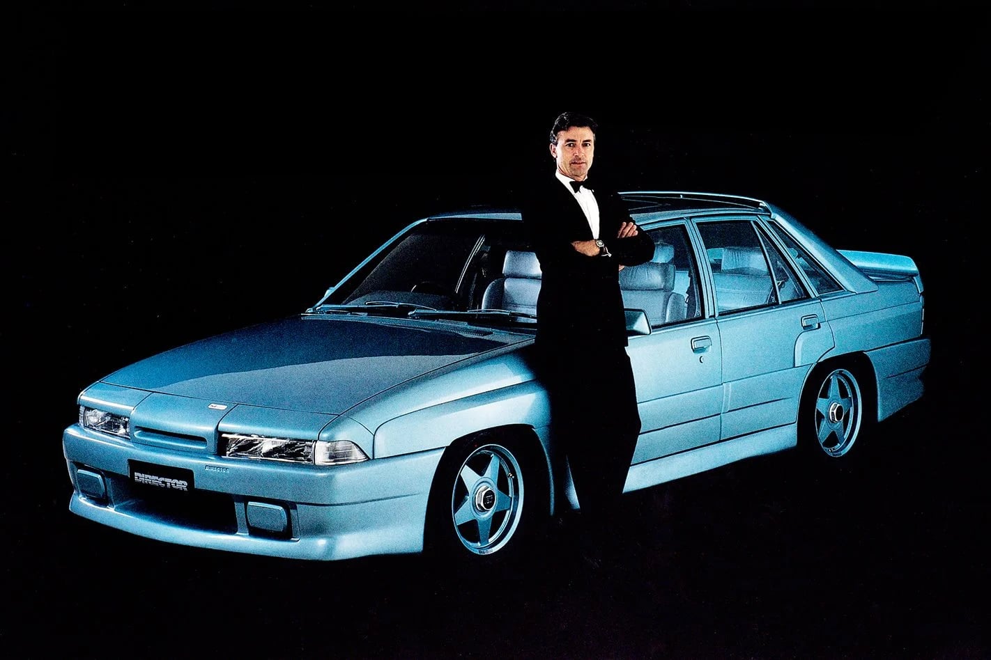 Man in a tuxedo standing with arms crossed beside a light blue vintage car against a black background