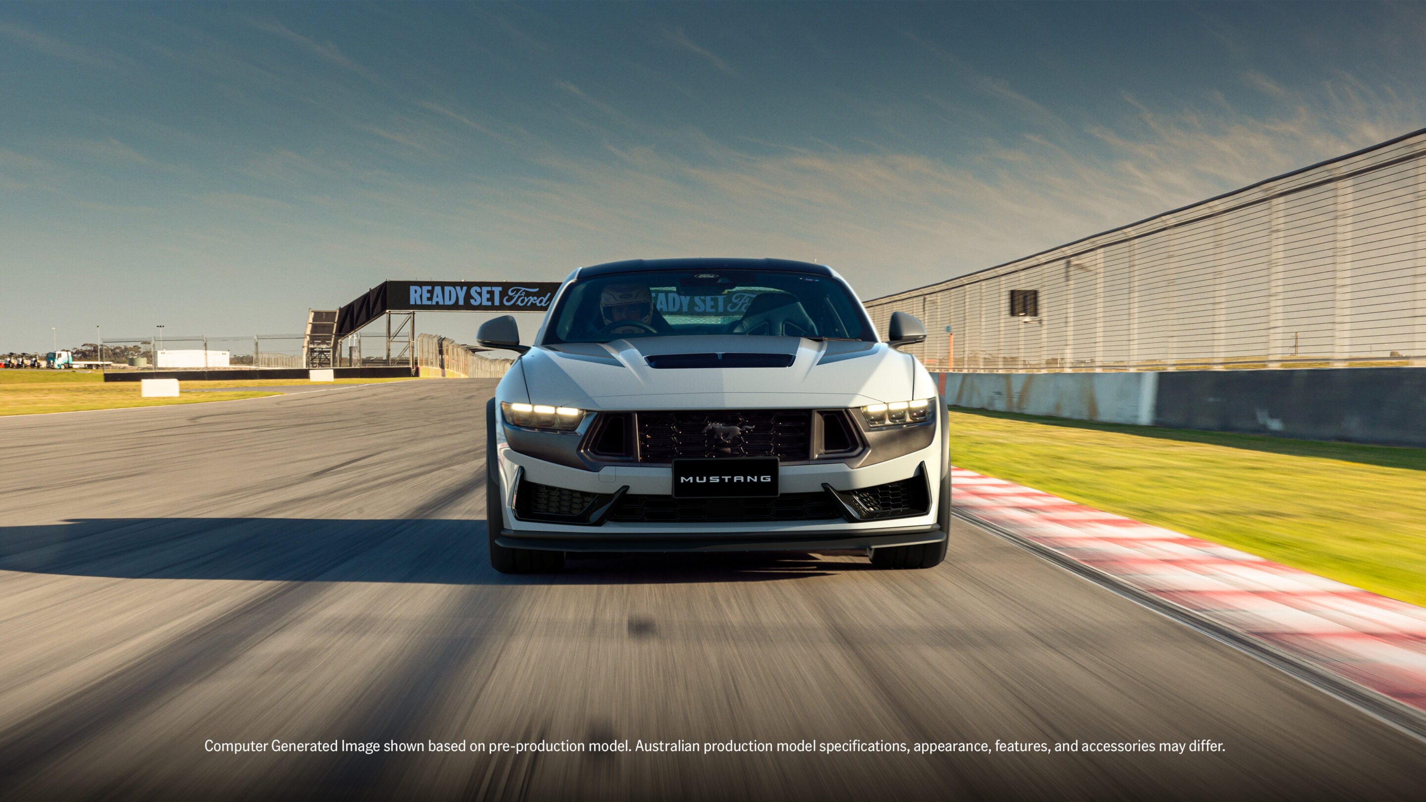 Front view of a white Ford Mustang speeding on a race track with motion blur and safety barriers in the background, Ready Set Ford banner visible behind the car
