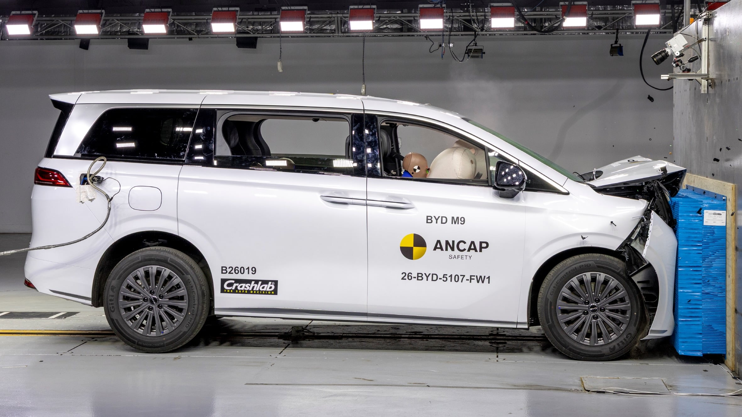 White BYD M9 crash-test van with a dummy in the driver seat inside a lab, ANCAP safety logo on the door.