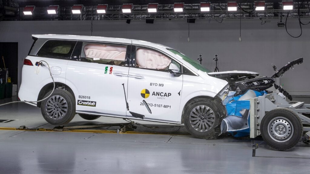 White crash-test minivan with deployed airbags inside a laboratory, ANCAP safety branding and damage to the front end visible, hood open.