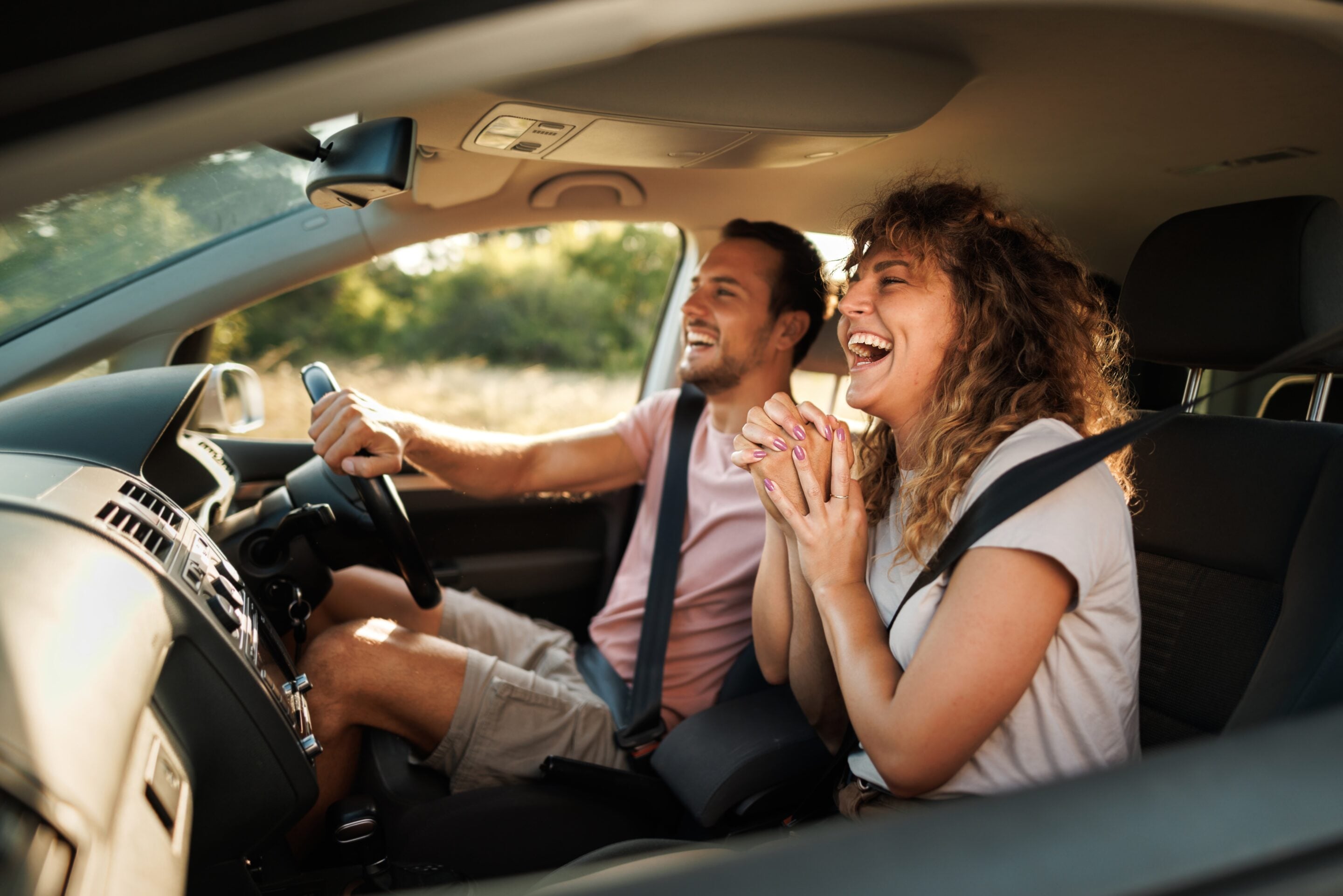 Two friends or a couple laugh together in a car, wearing seatbelts, while the driver holds the wheel and chats joyfully.
