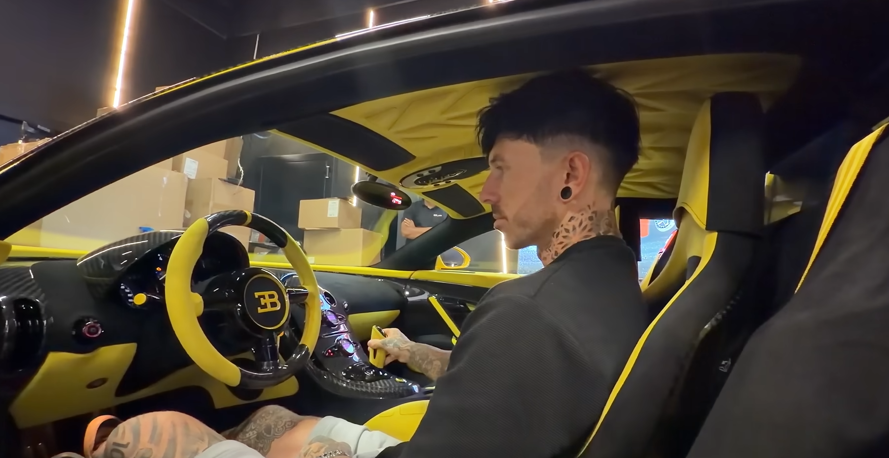 Man with tattoos sits in the driver’s seat of a yellow-and-black luxury car, with carbon-fiber dash and bright accents visible.