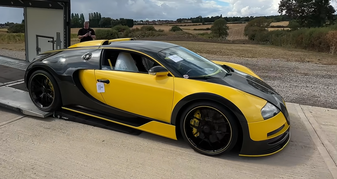 Two-tone yellow and black Bugatti Veyron parked on a car transporter ramp outdoors, with a rural field backdrop.