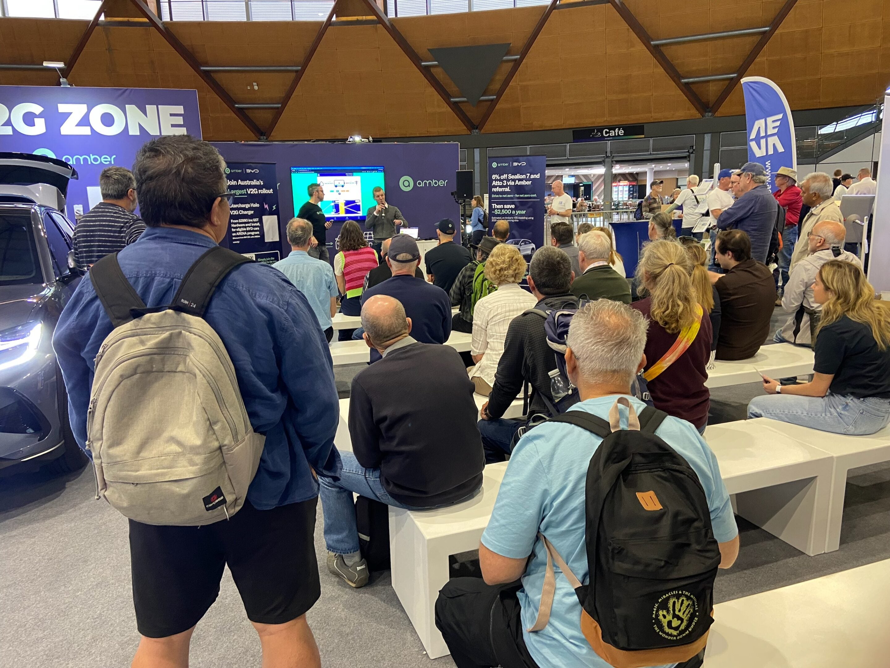 Audience at a tech expo watches a presenter on a stage with large screens and a purple booth backdrop.
