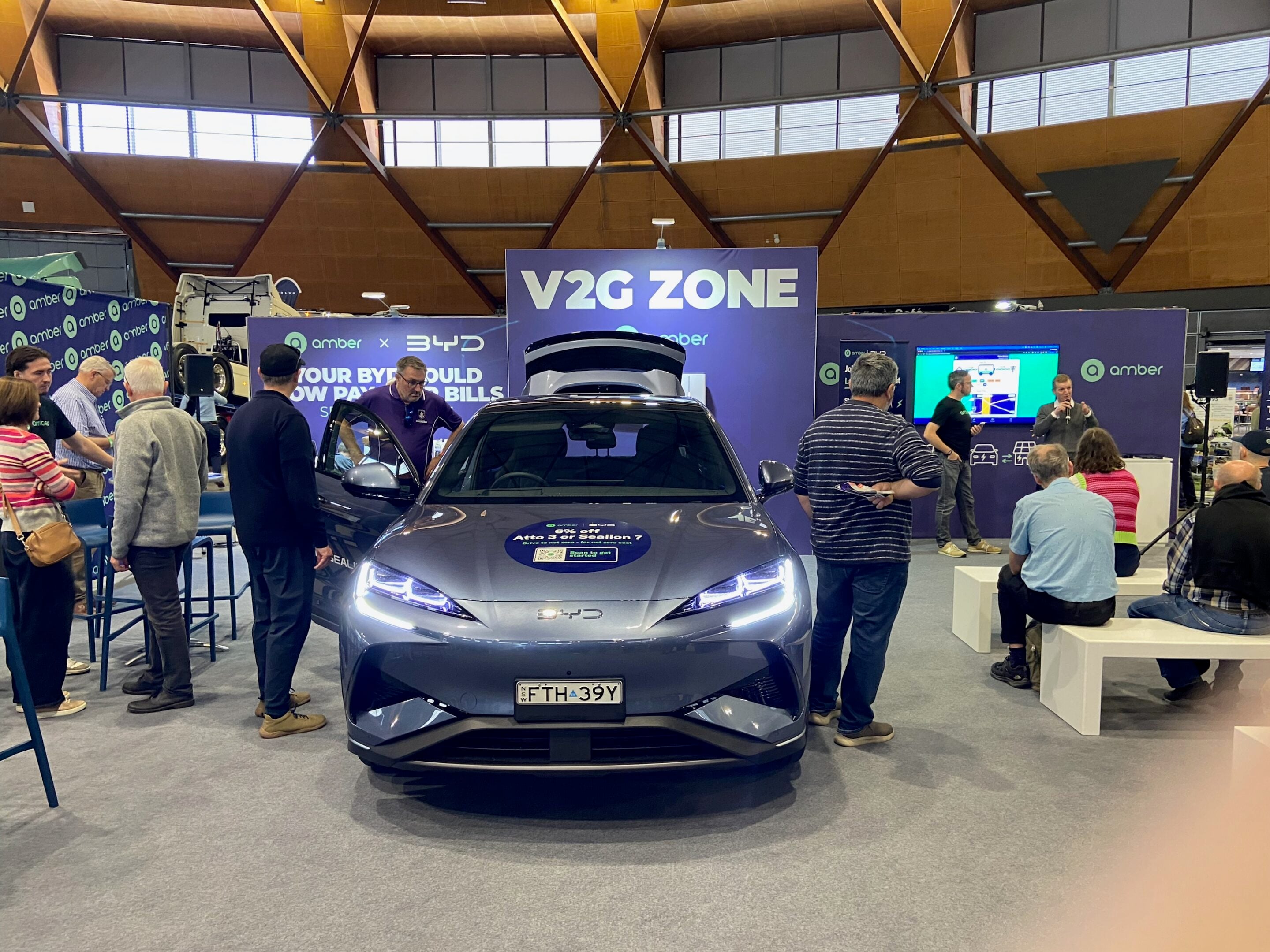 Blue car on a showroom floor at an exhibition with a large purple banner reading 'V2G ZONE' and people gathered around the vehicle's front.