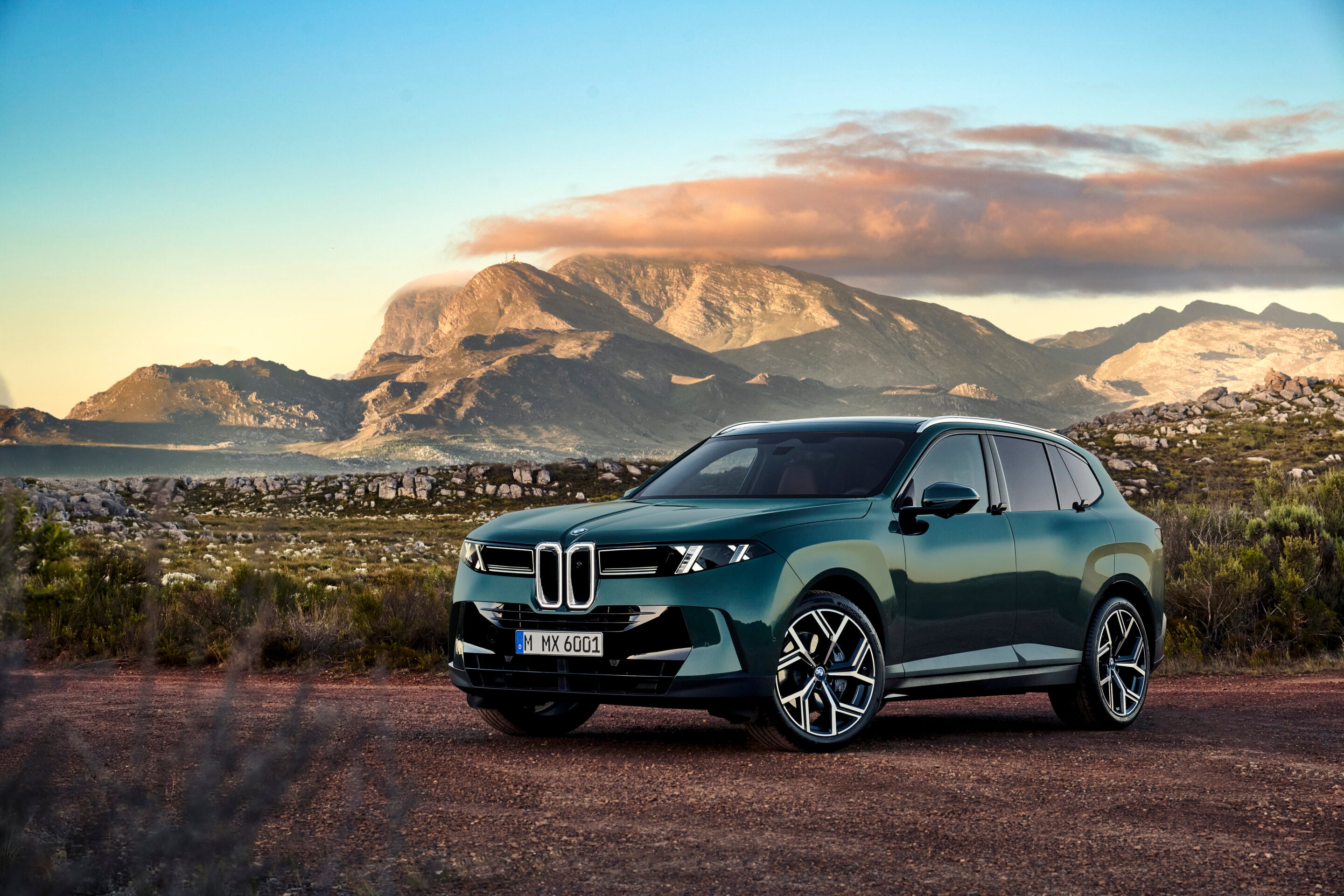 Teal SUV parked on a dirt road with rugged mountains and a blue sky in the background.