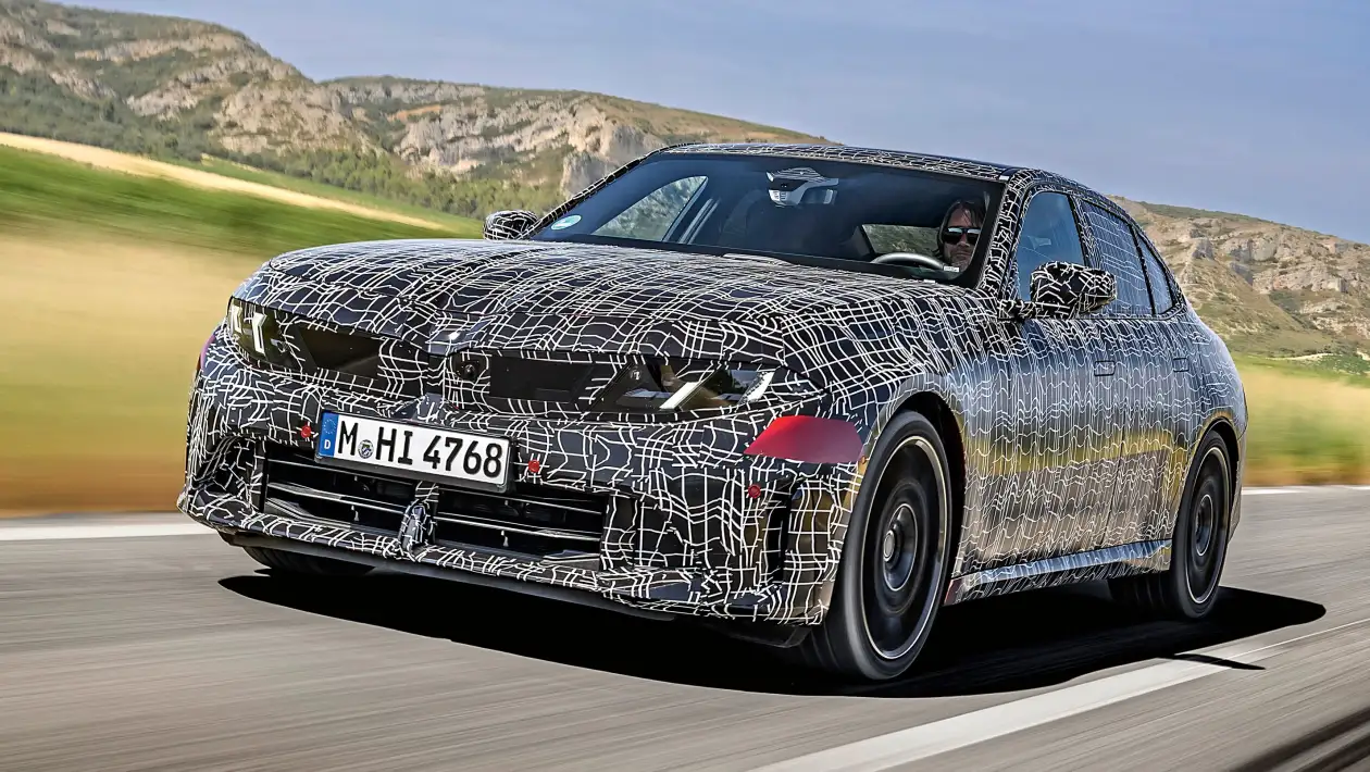 Camouflaged sedan prototype driving on a road in a rural landscape, with a white web-like wrap hiding its design details