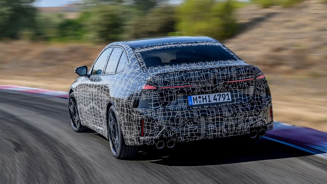 Camouflaged sedan prototype driving on a race track, rear view, white abstract wrap hides details of the car's design.
