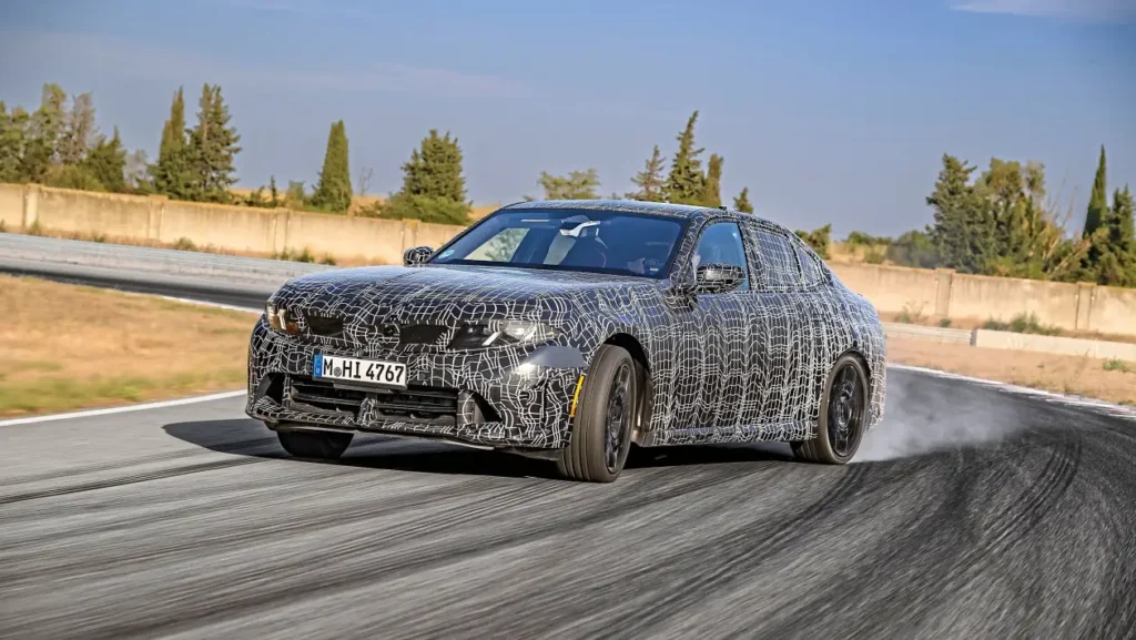 Camouflaged prototype sedan taking a wide turn on a race track, with smoke at the rear tires.
