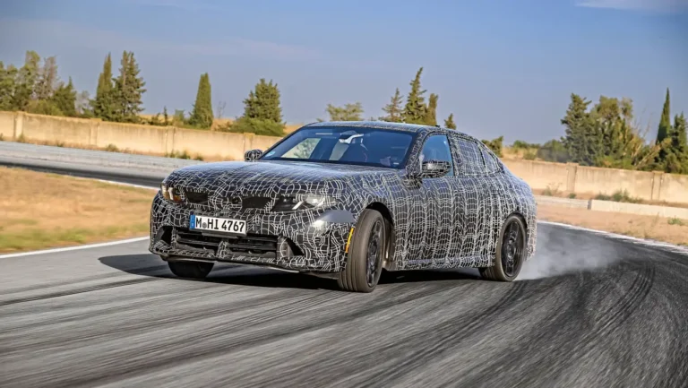 Camouflaged prototype sedan taking a wide turn on a race track, with smoke at the rear tires.