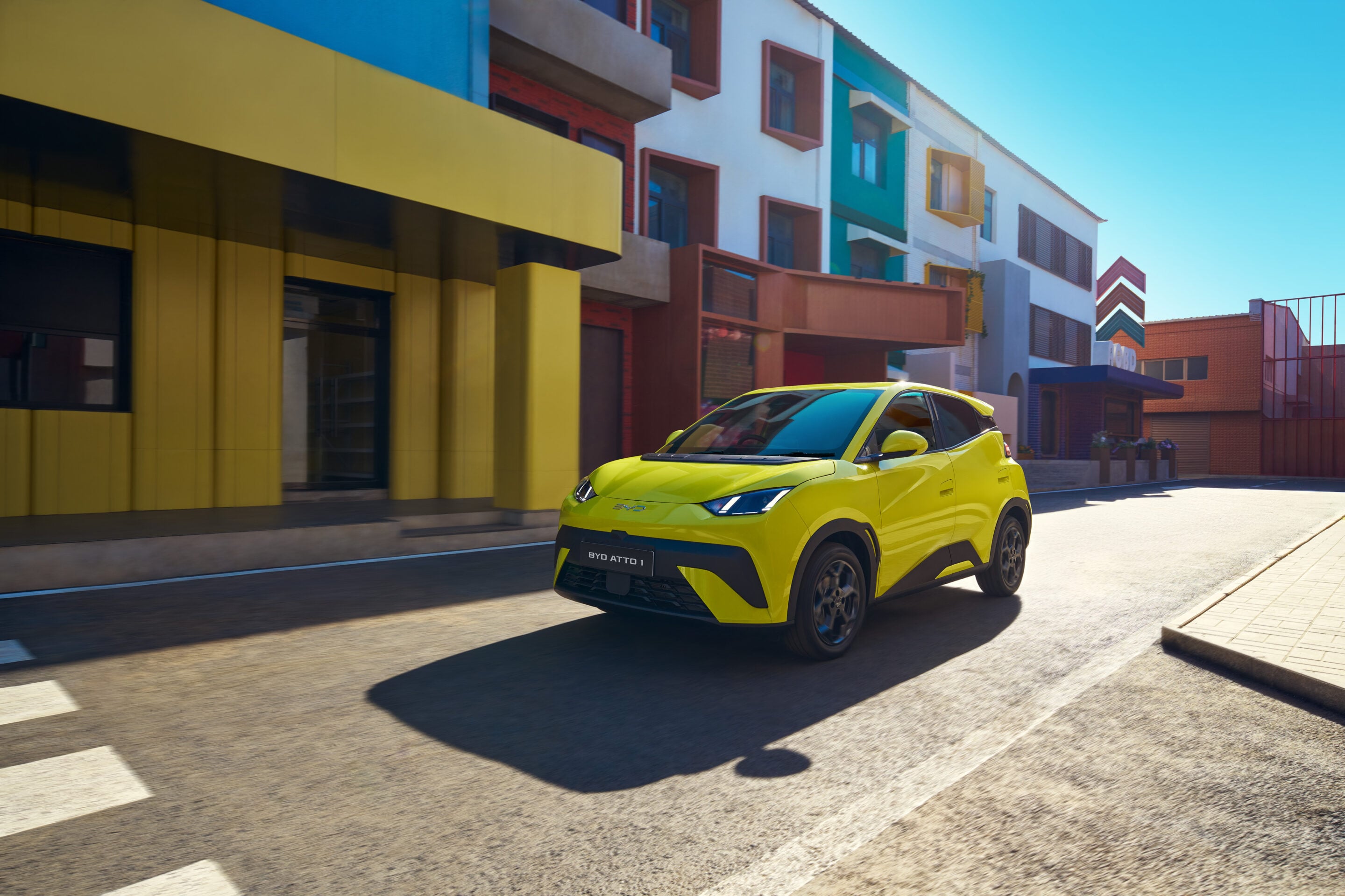 Bright yellow BYD ATTO 3 SUV driving along a sunny city street with colorful modern buildings in the background.