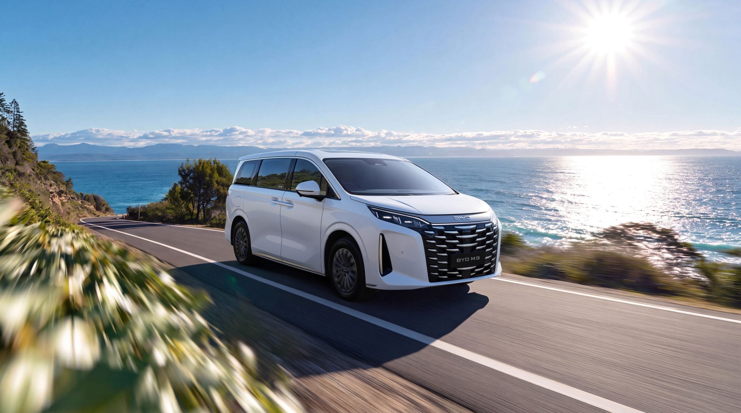 White BYD M9 minivan driving along a coastal highway with the ocean on the right under a bright sun.