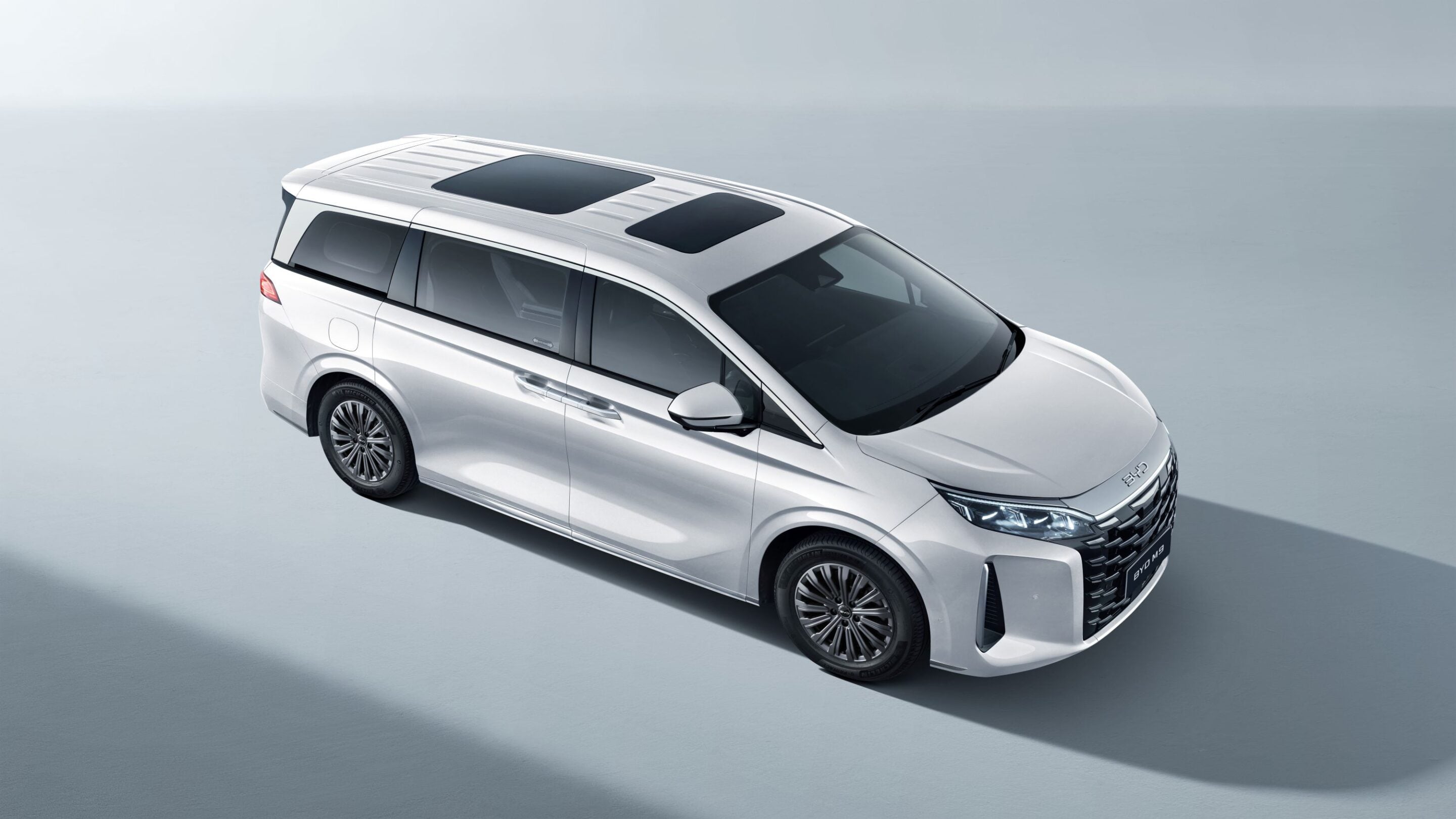 Top-down view of a white luxury minivan with two sunroofs and a sleek front grille against a pale background.