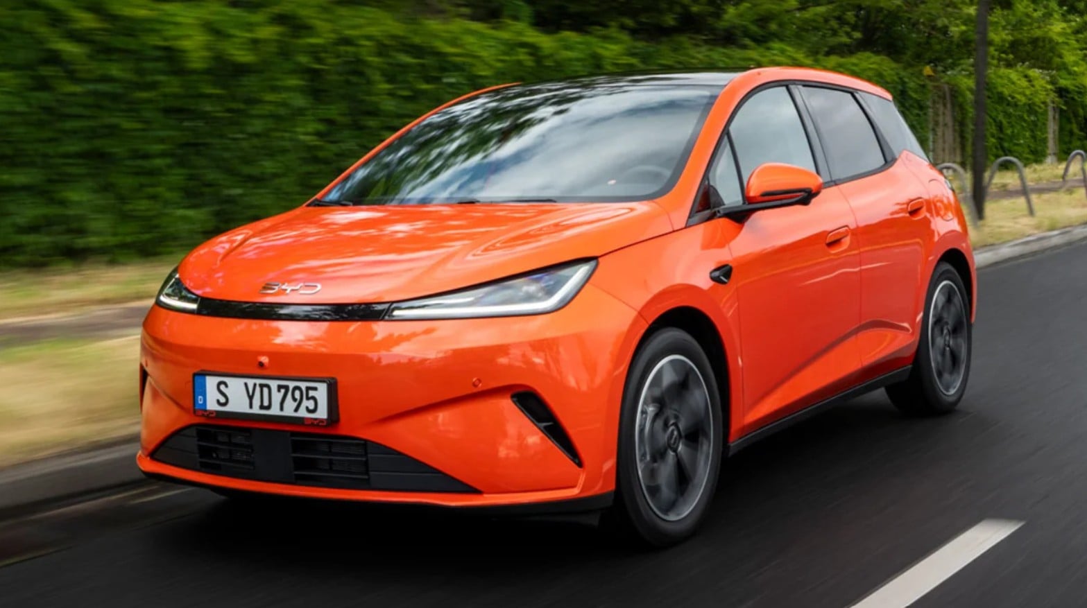 Orange compact hatchback car driving along a suburban road, front three-quarter view, motion blur in the background