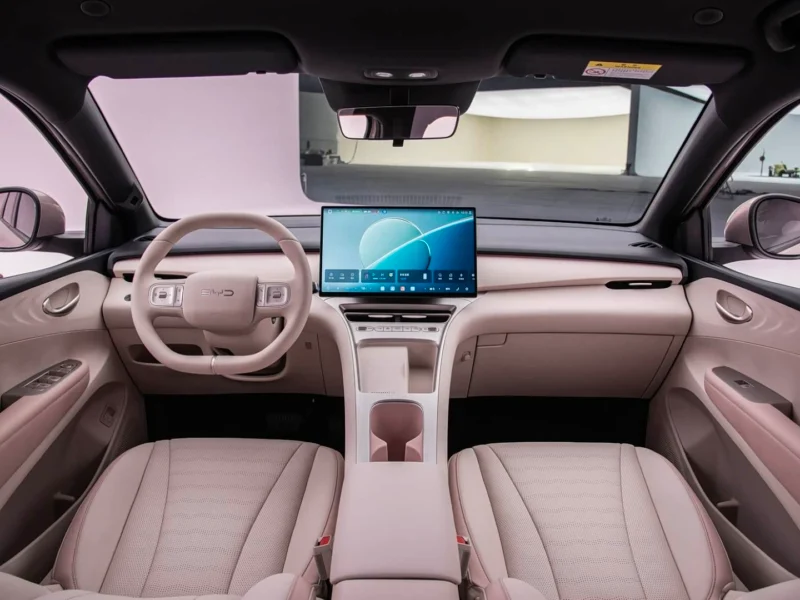 Beige luxury car interior with a large central touchscreen, steering wheel on the left, and front seats visible.