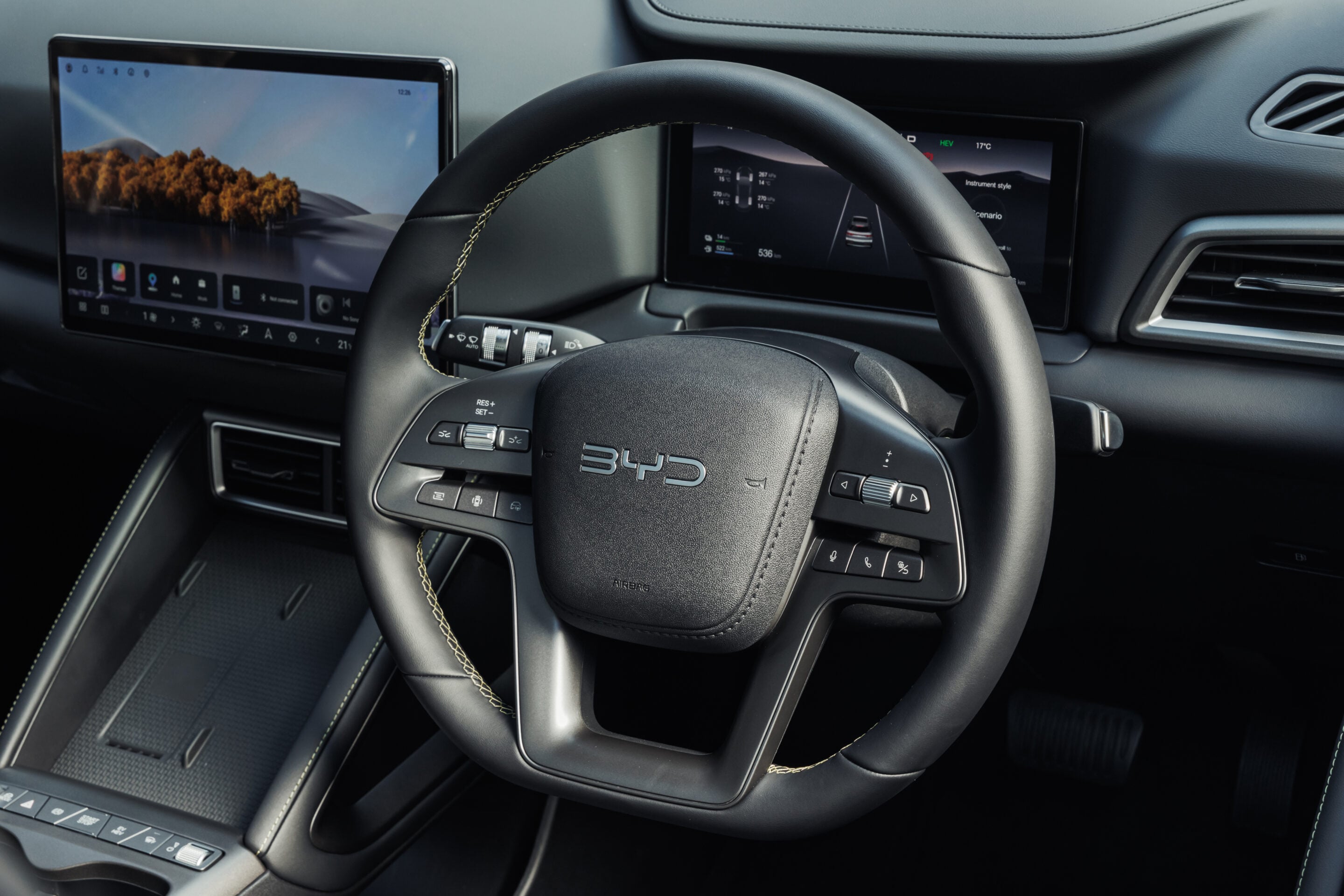 Steering wheel in a modern car cockpit with two large screens: a left tablet-style display showing a landscape wallpaper and a right digital instrument panel showing vehicle data.