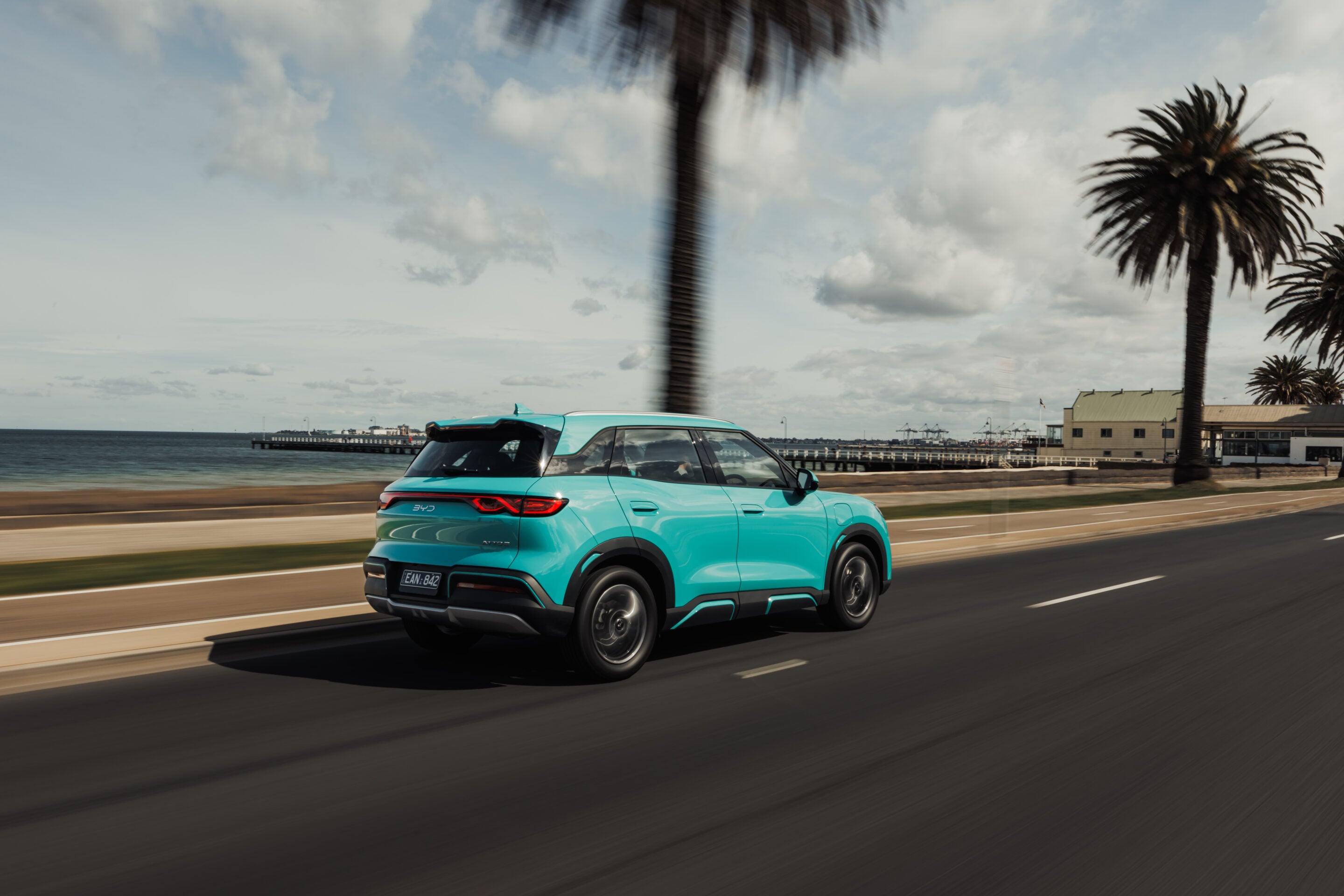 Turquoise SUV driving along a coastal road with palm trees and a pier in the background.