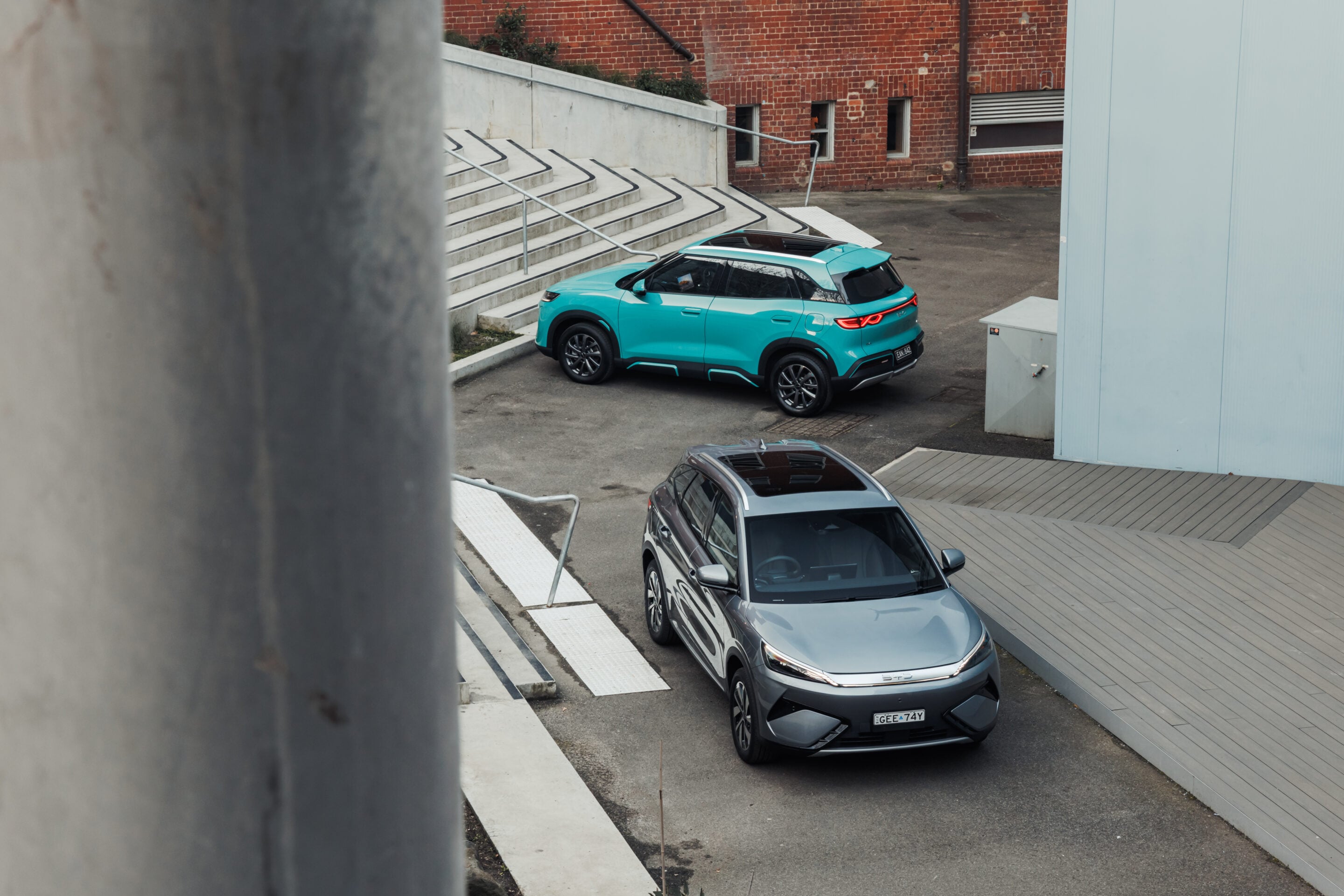 Rear three-quarter view of a turquoise SUV parked in a concrete parking area near a brick building with stairs and railings in the background
