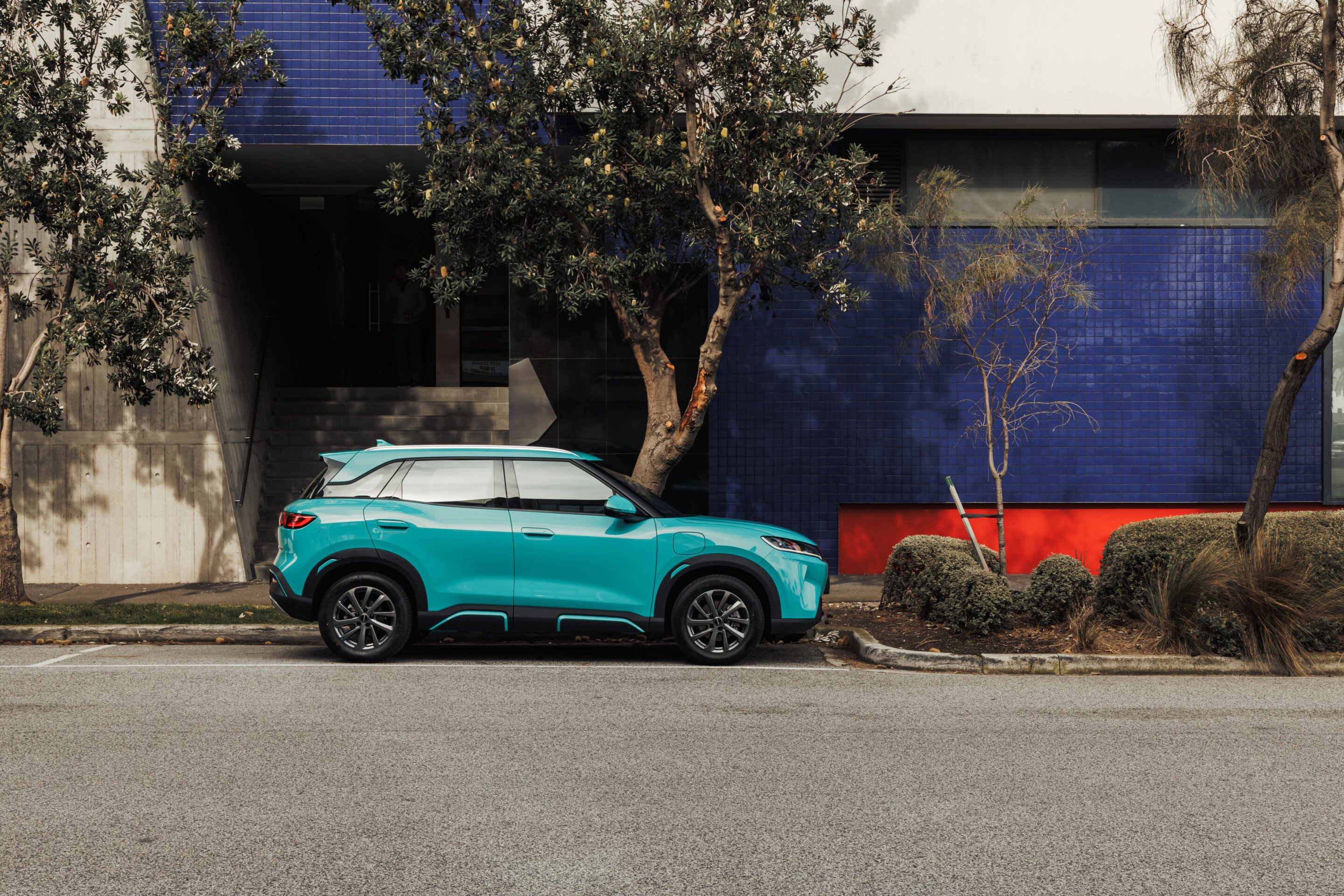 Turquoise SUV parked along a curb in front of a blue-tiled wall, with trees partially obscuring the building.