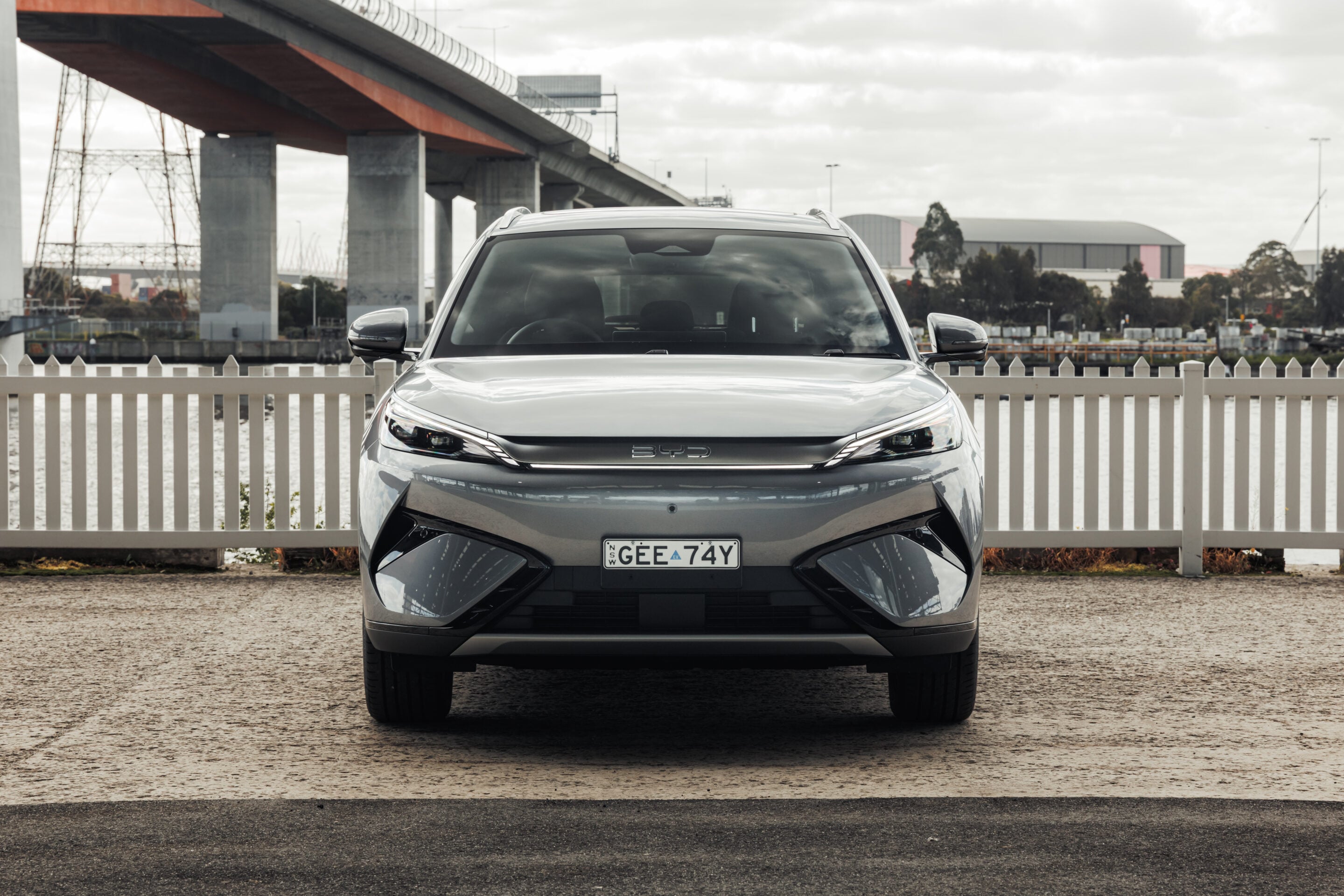 Front view of a silver SUV parked behind a white picket fence with an elevated road/bridge in the background.