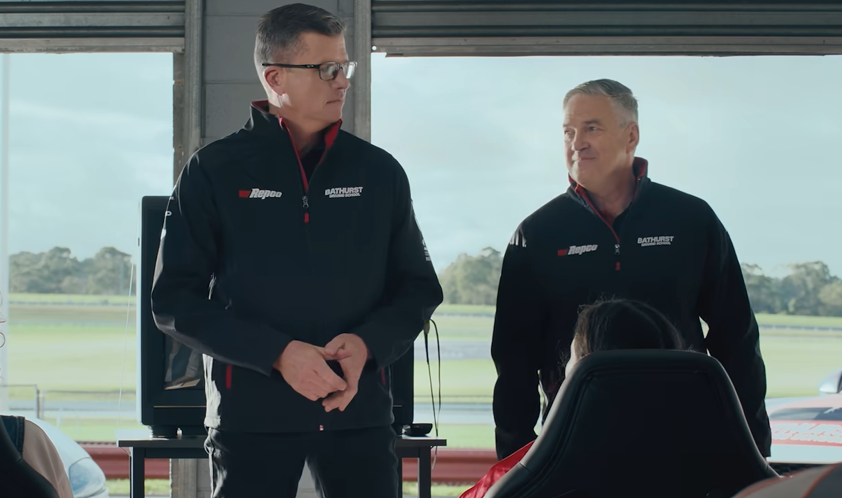 Two men in black jackets with red zippers stand and speak in a garage overlooking a racetrack.