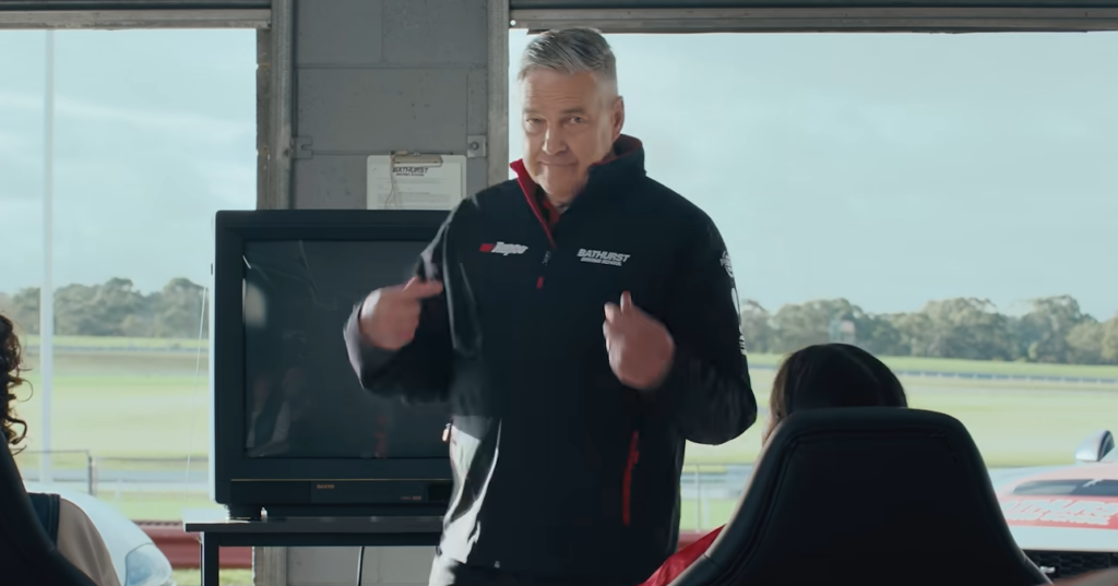 Man in a Bathurst-branded jacket speaks to an audience in a pit-area room, standing in front of a TV monitor.