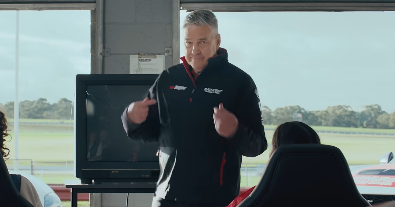 Man in a Bathurst-branded jacket speaks to an audience in a pit-area room, standing in front of a TV monitor.