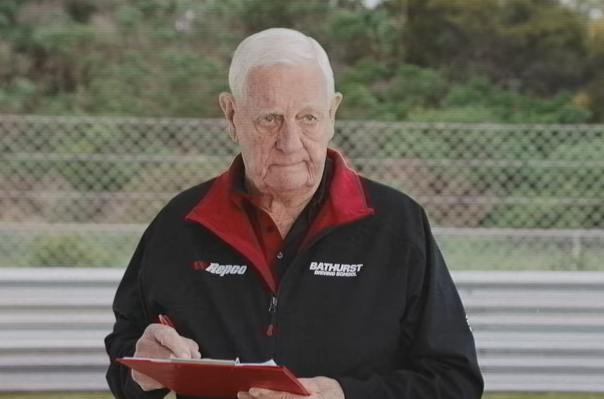 Older man with white hair wearing a black jacket with red lining, writing on a red clipboard at a race track behind a chain-link fence.