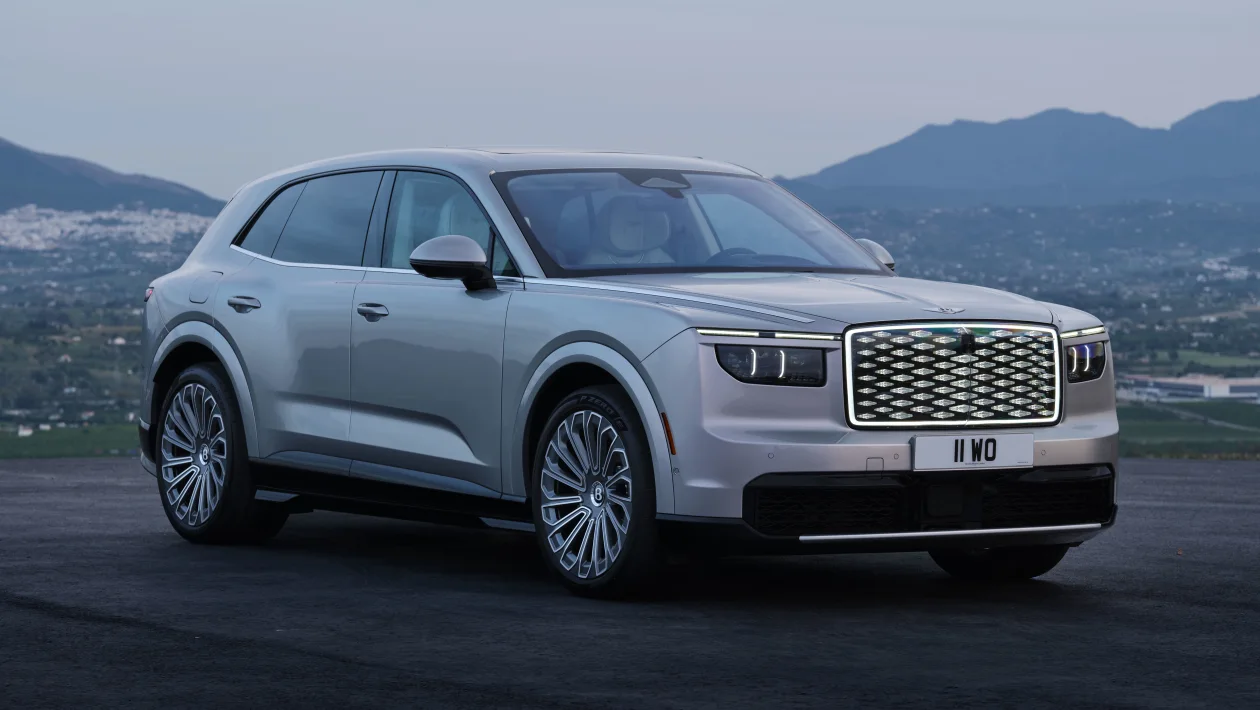 Three-quarter front view of a silver luxury SUV parked on a dark asphalt surface with a mountainous landscape in the background, emphasizing its bold grille and LED headlights.