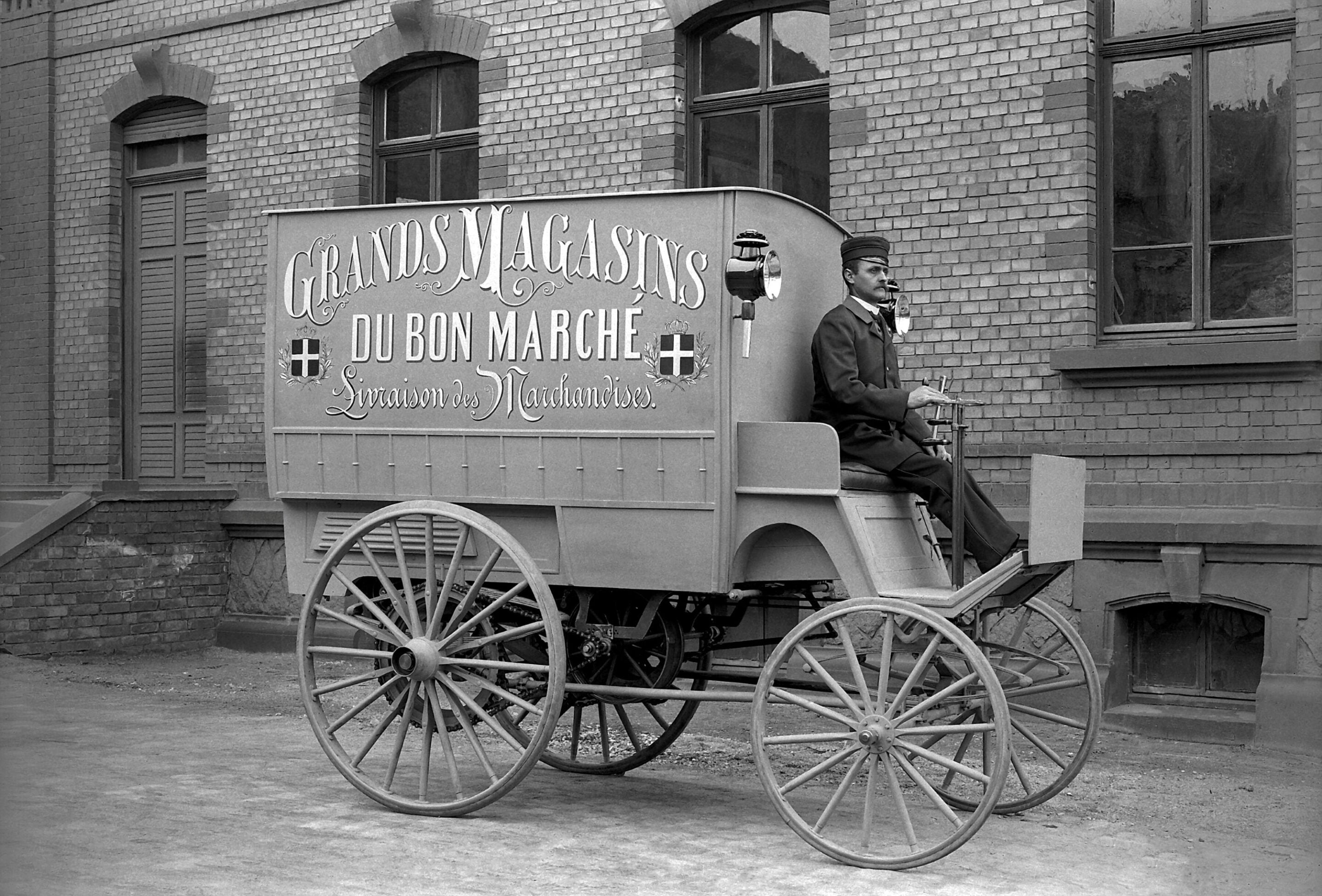 Black-and-white photo of a uniformed driver on a vintage delivery wagon in front of a brick building; wagon sign reads Grands Magasins du Bon Marché.