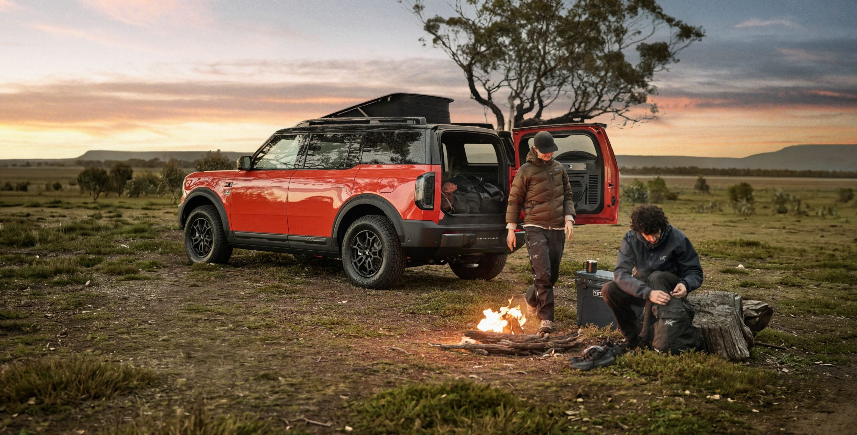 Red off-road SUV with rooftop tent and open rear, two campers by a small campfire at sunset in a wide open field.