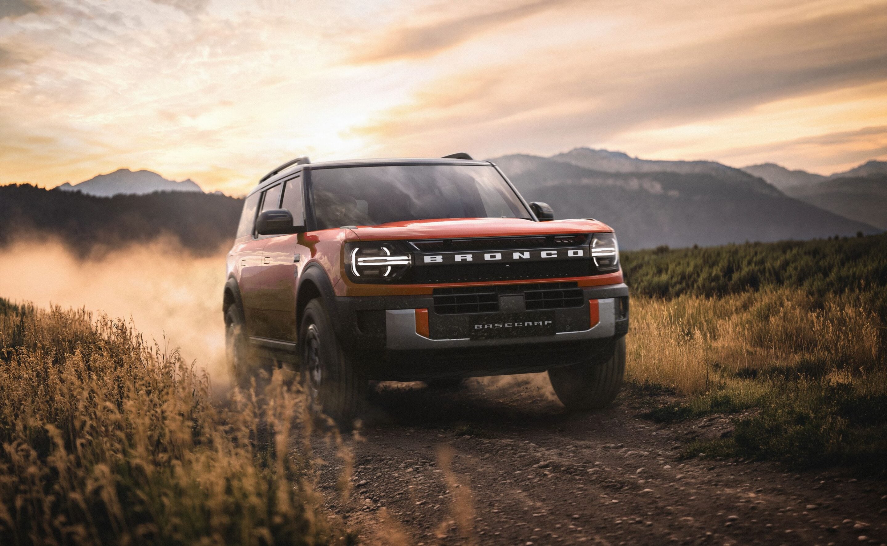 Red Ford Bronco driving through a dusty field at sunset with mountains in the background.
