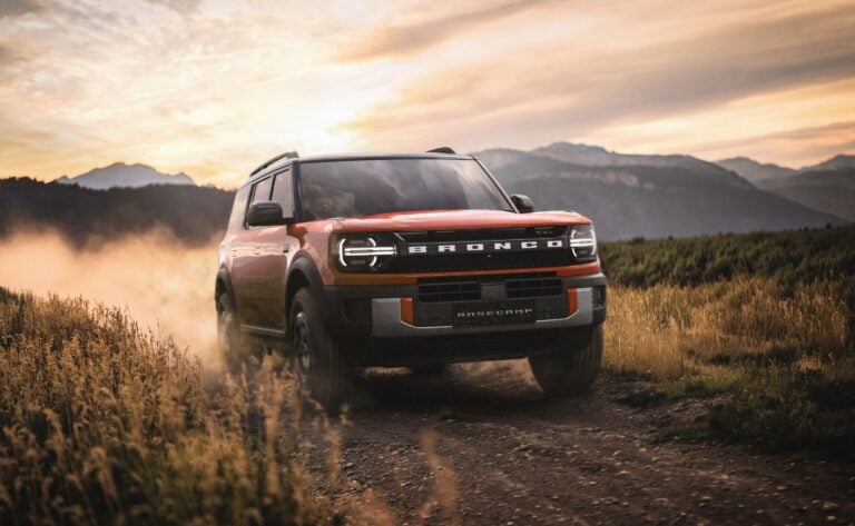 Red Ford Bronco driving through a dusty field at sunset with mountains in the background.
