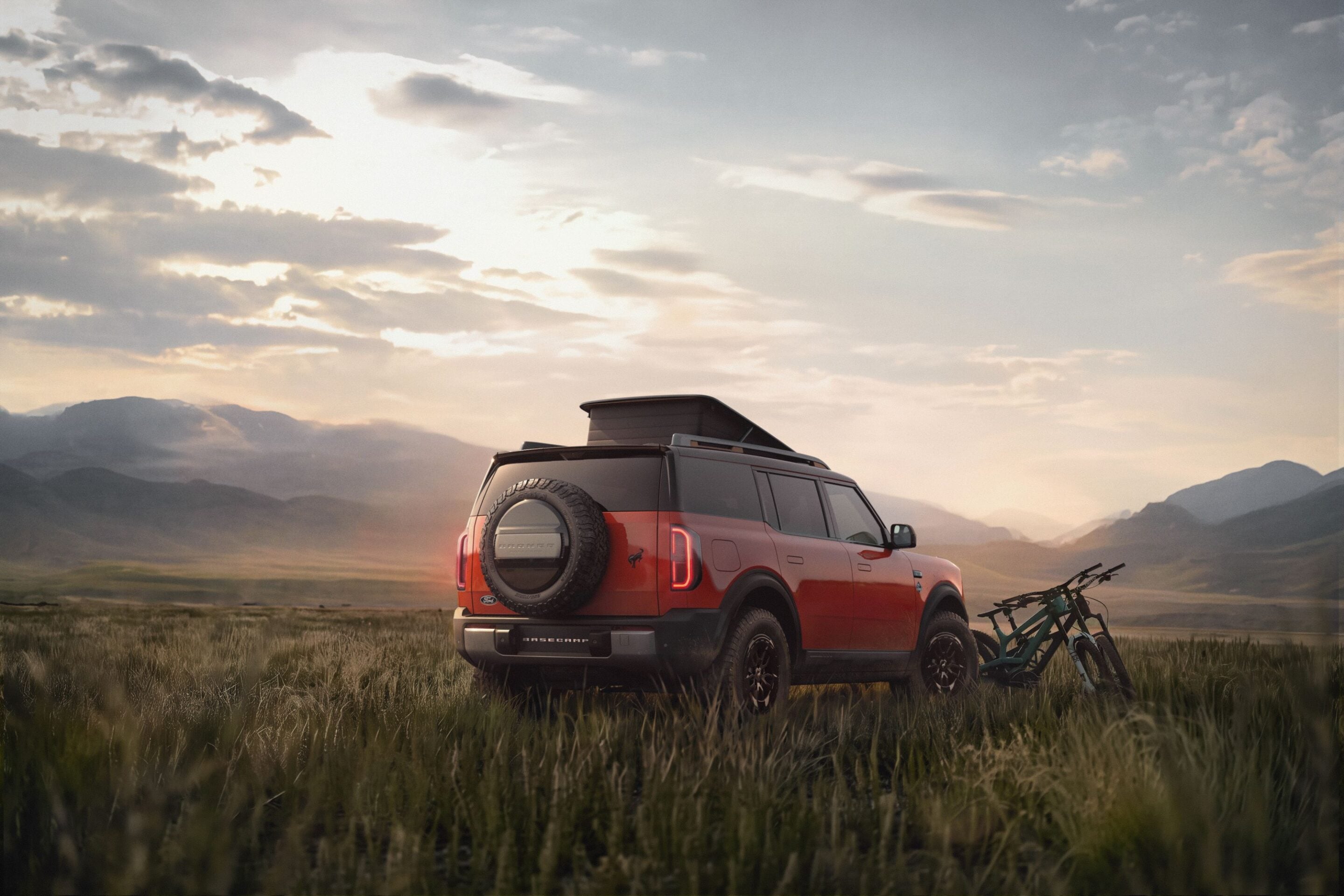 Orange SUV with a mounted spare tire on the back, parked in tall grass with mountains and a sunset sky in the background, bikes attached nearby.