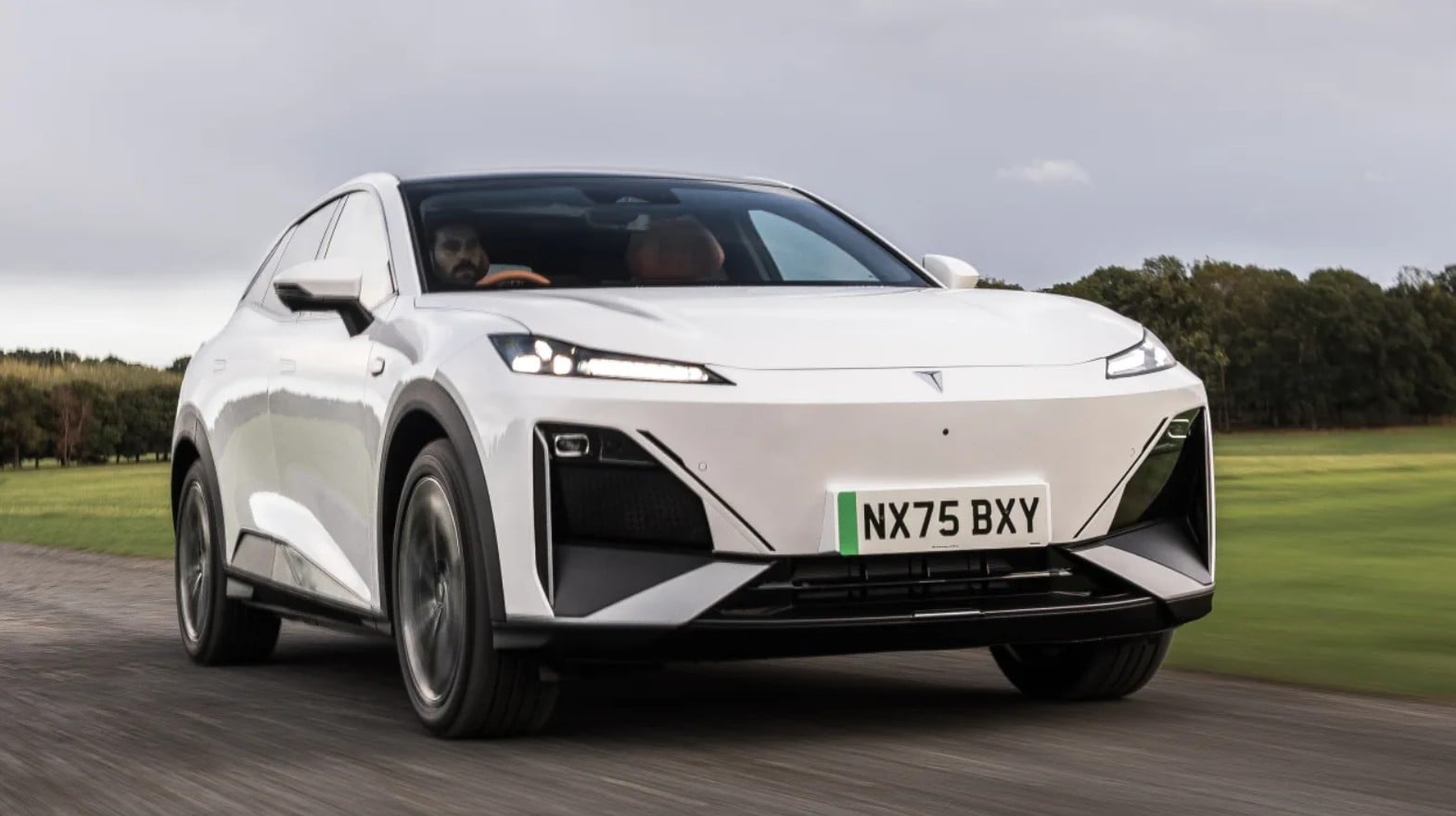 White futuristic-looking electric SUV driving on a road with grass and trees in the background; license plate NX75 BXY visible on the front.