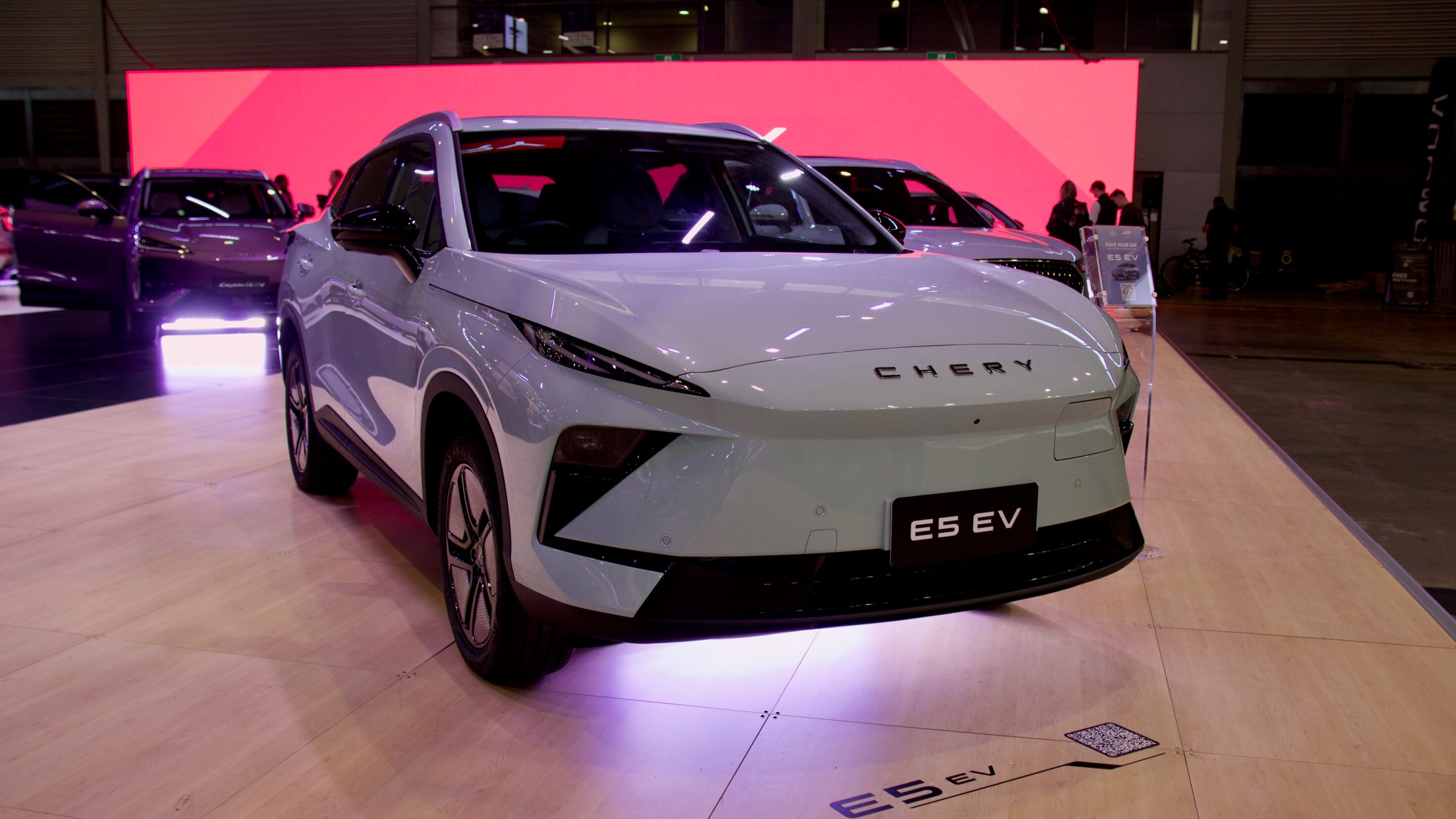 Front view of a white Chery E5 EV on a showroom display with a bright pink backdrop behind it.