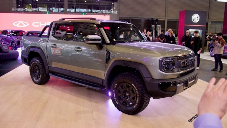 Three-quarter front view of a silver pickup on display at a car show with visitors in the background taking photos.