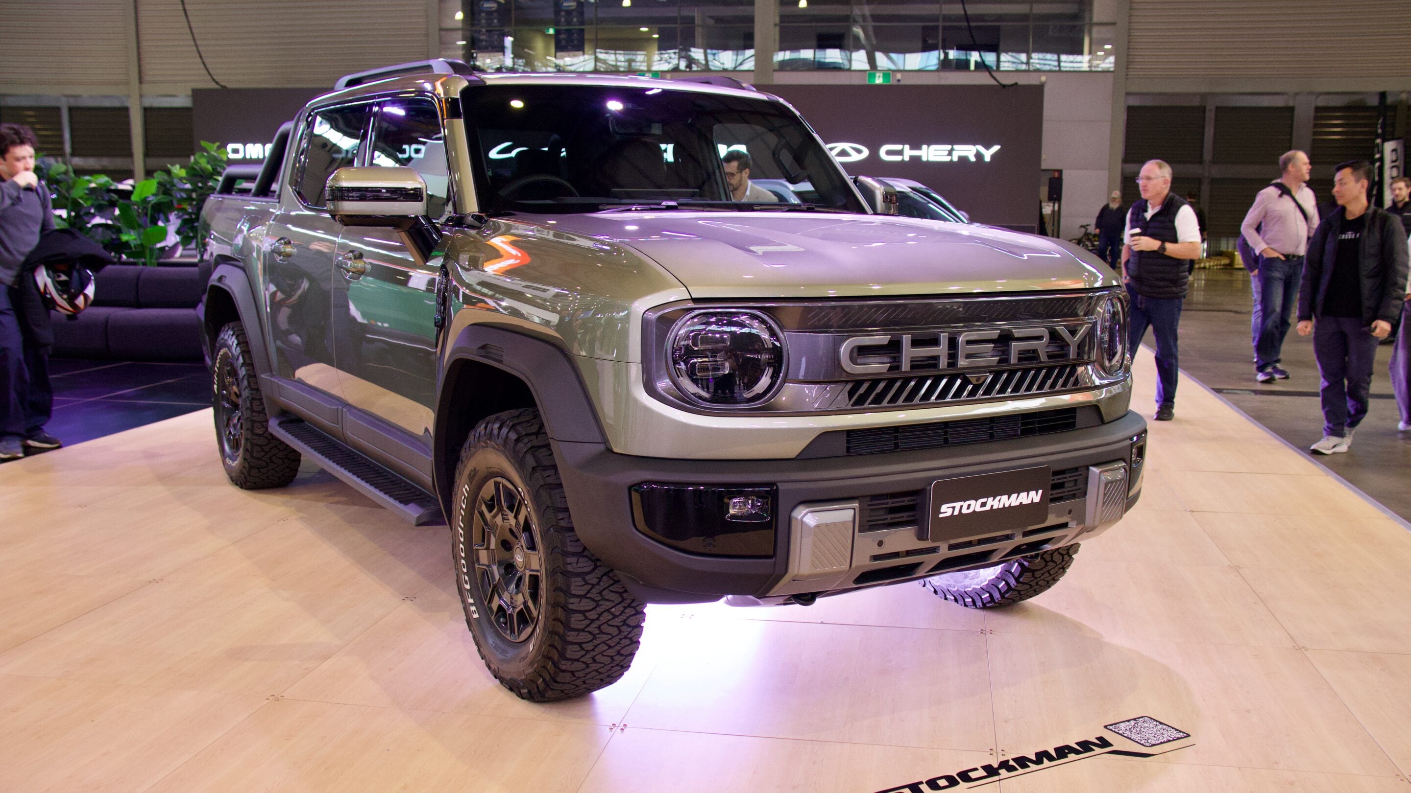 Silver pickup truck on a showroom platform with a CHERY badge and Stockman plate, attendees in the background at an auto show.