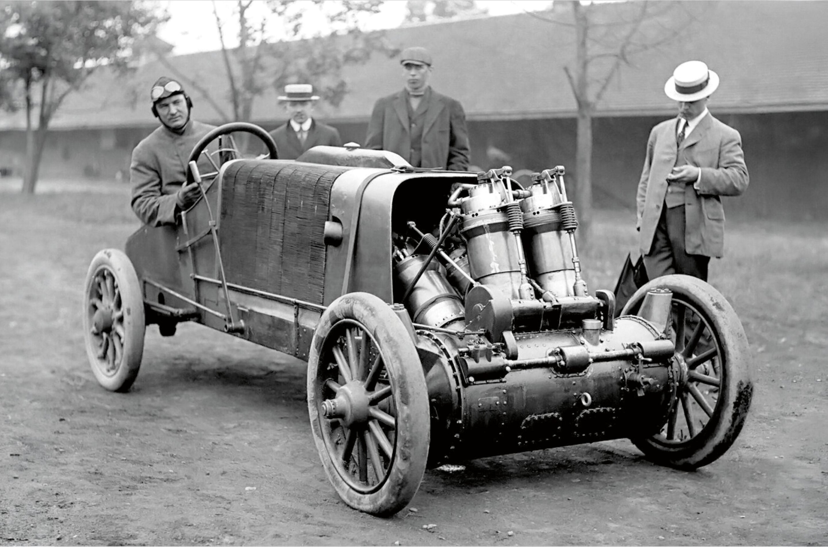 Vintage open-top race car with exposed engine, attended by three men in early 20th-century attire and driving goggles and caps.