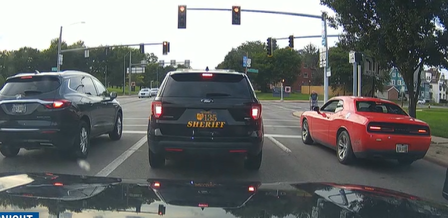 Three cars at an intersection: a gray SUV on the left, a black sheriff's SUV in the middle with 'SHERIFF' in gold letters, and a red sports car on the right; traffic lights overhead.