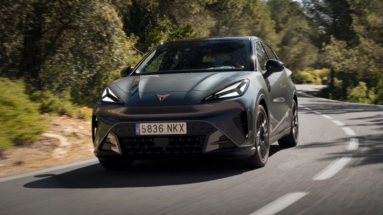 Front-three-quarter view of a gray hatchback driving along a tree-lined road, showing its sporty front end and angled headlights.