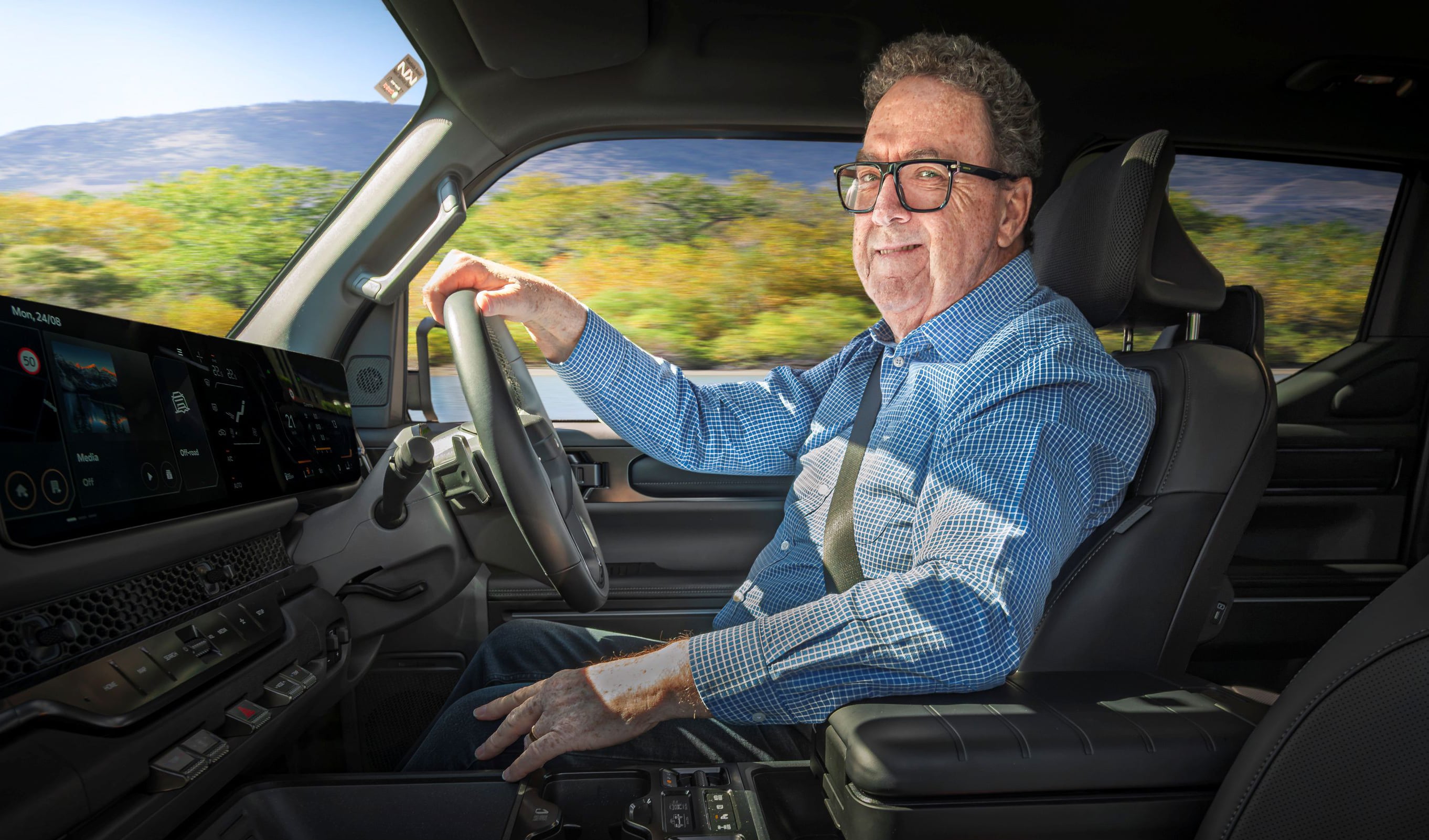 Older man wearing glasses in a blue checkered shirt driving a car, with landscape visible outside the window.