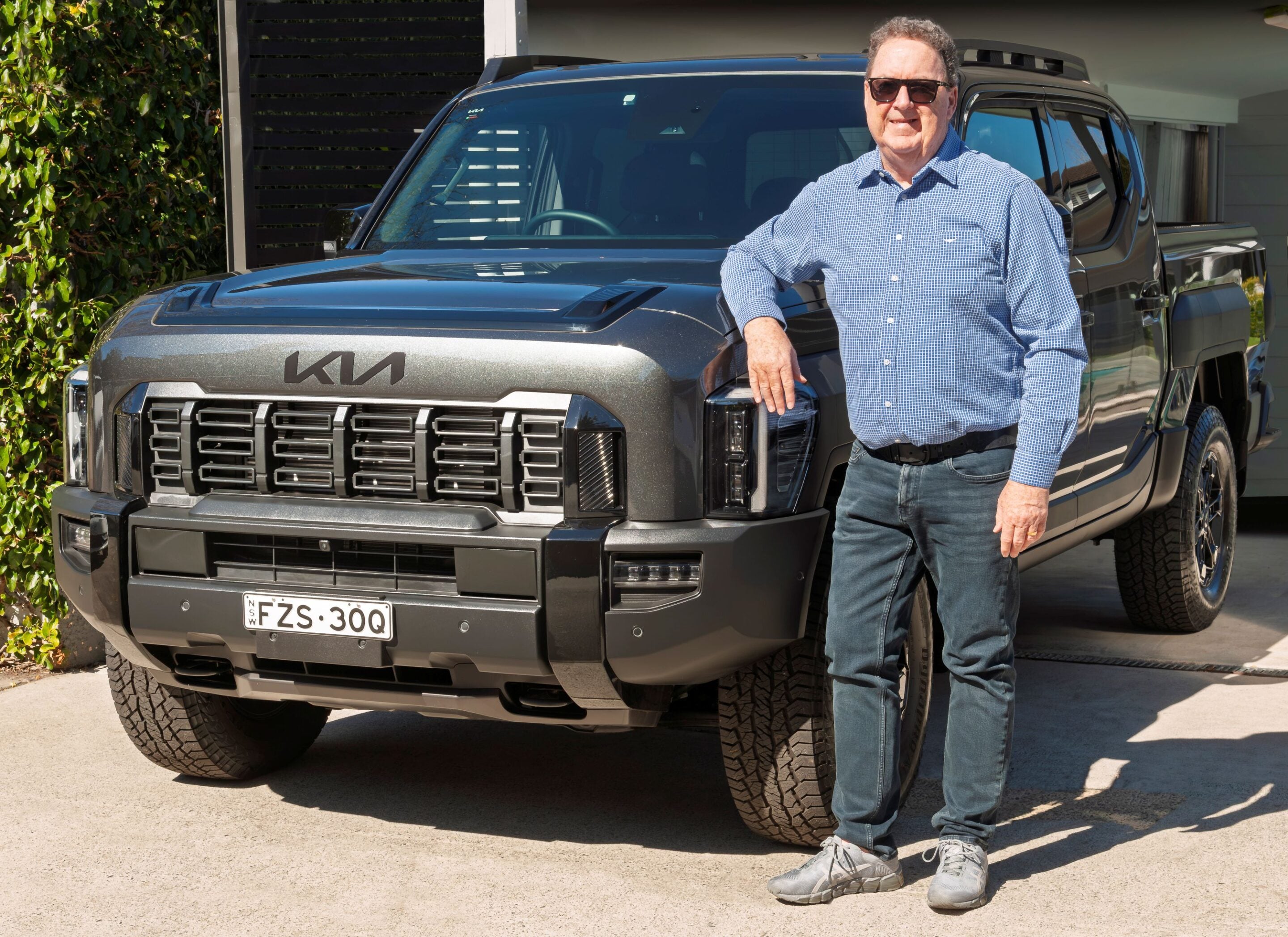 Older man in a blue checkered shirt and sunglasses leaning on a gray Kia pickup truck in a driveway.