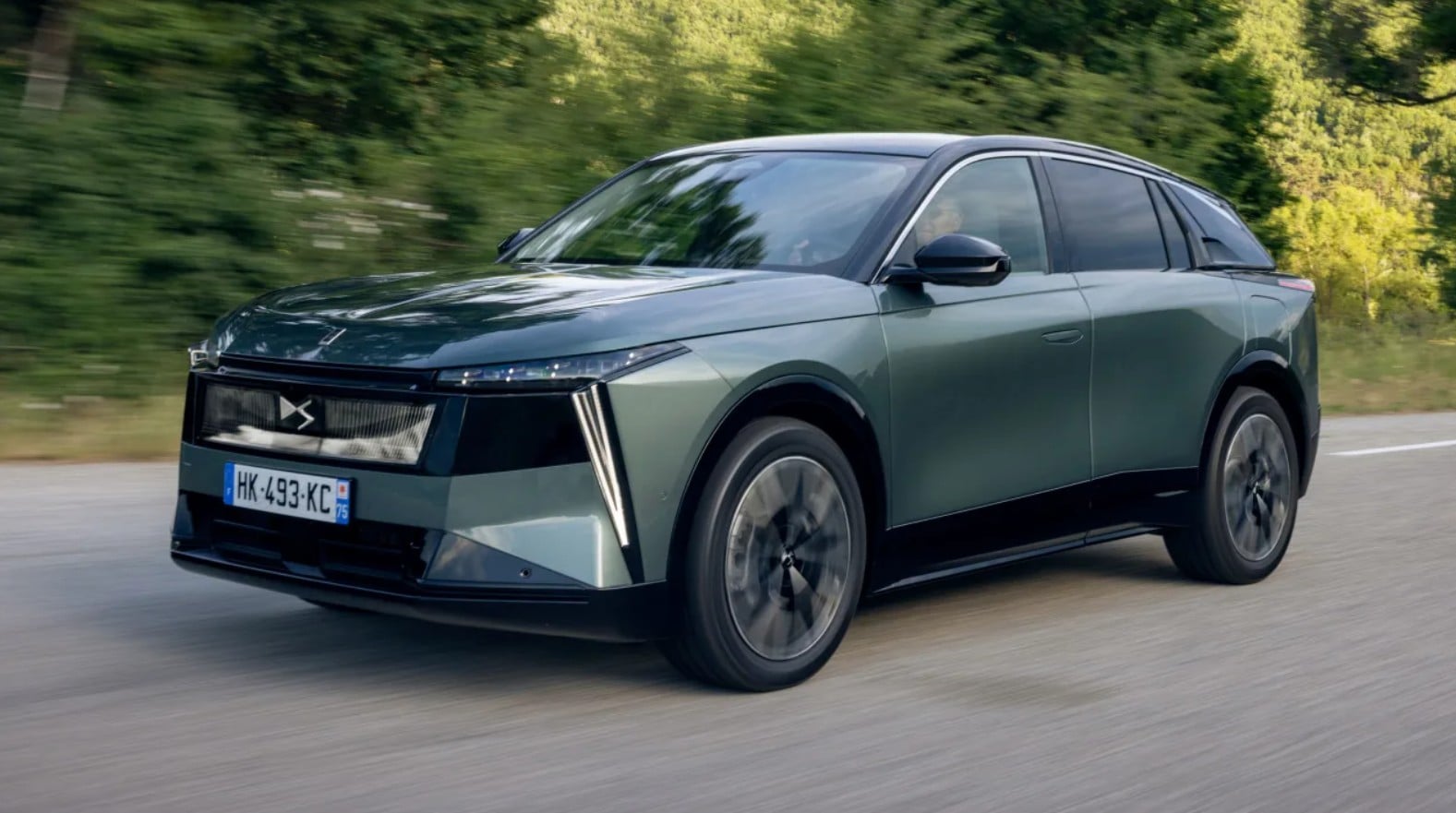 Front three-quarter view of a metallic green SUV driving on a road with a blurred tree-lined background, motion blur showing speed.