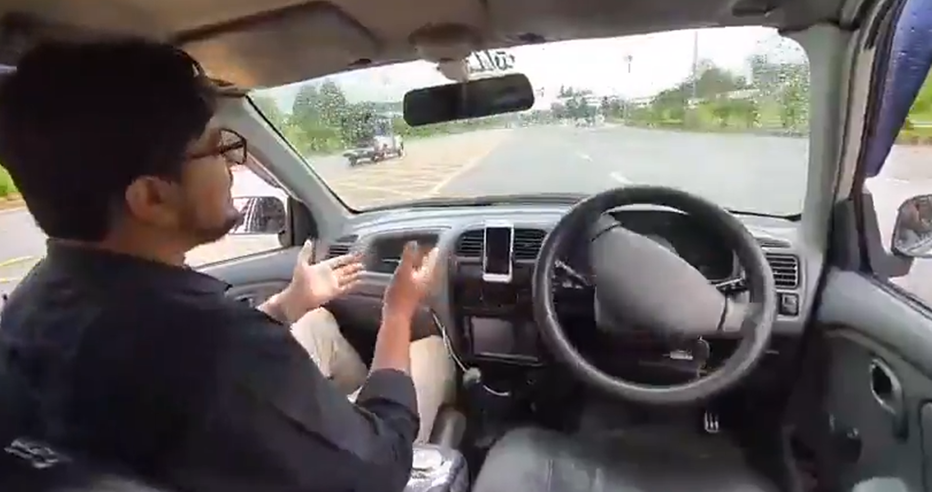 Man wearing glasses sits in the driver’s seat, speaking with animated hand gestures as he drives, road ahead visible through the windshield.