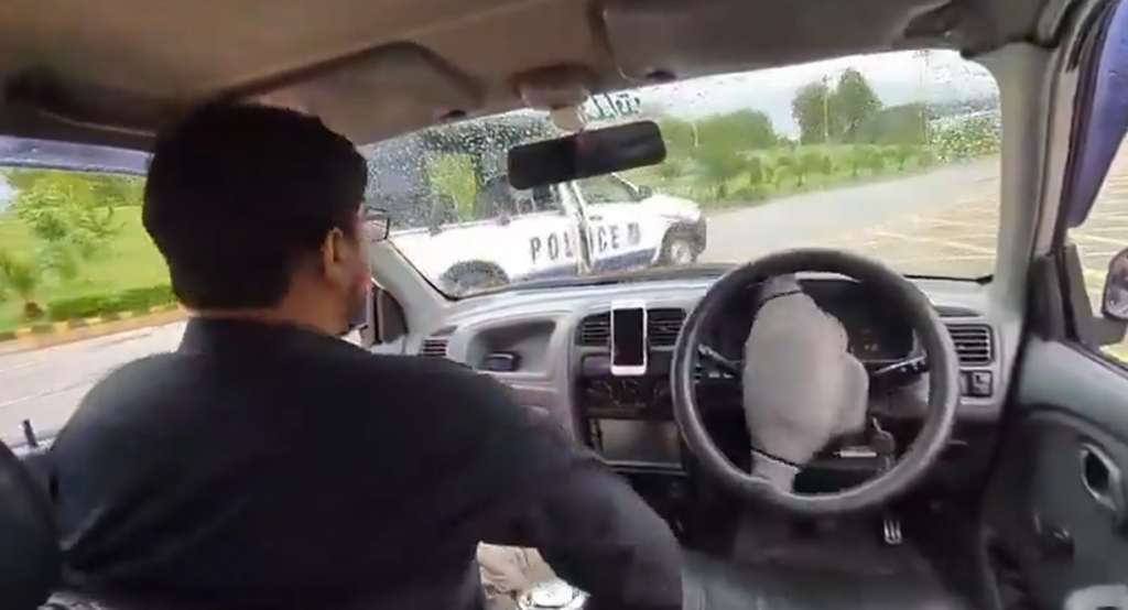 Interior of a patrol car with the driver in foreground; a police SUV is visible ahead through a rain-spattered windshield.