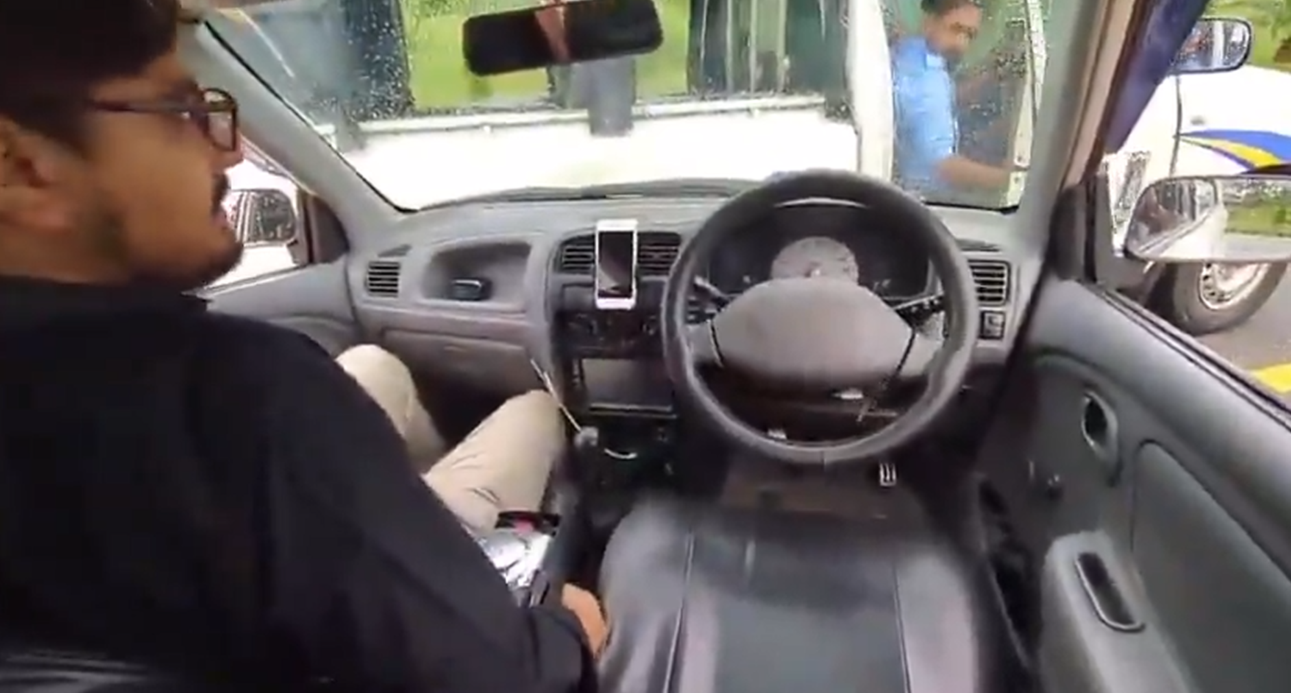Inside a car, a man wearing glasses sits in the driver’s seat with the steering wheel in front of him and a smartphone mounted on the dashboard, visible through the windshield.